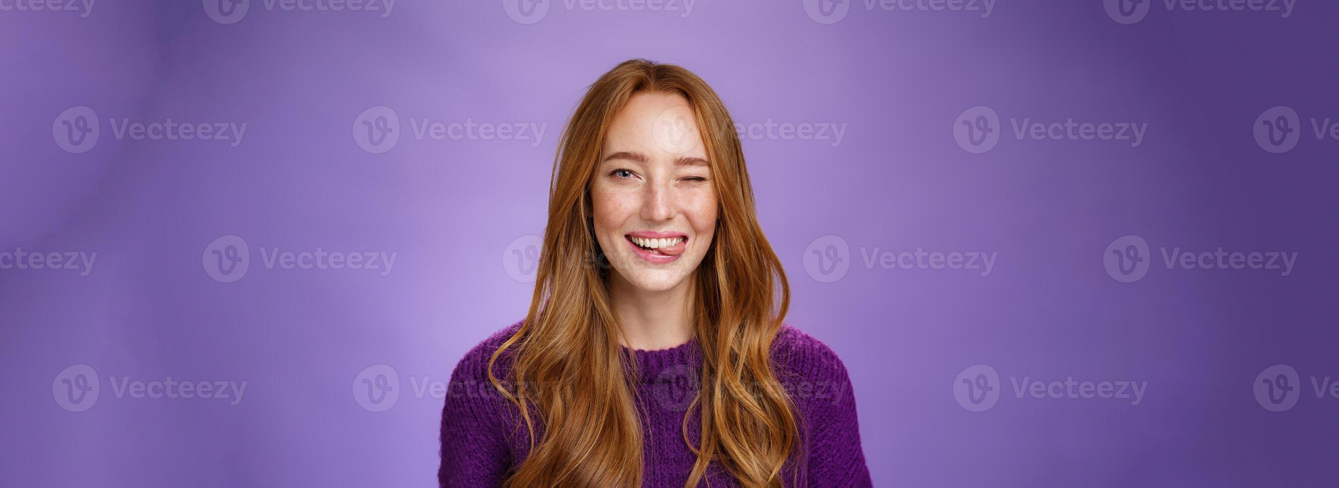 Close-up shot of sincere and happy funny cute redhead female with long ginger hair winking happily and sticking out tongue childish and carefree posing cheerful over purple background photo