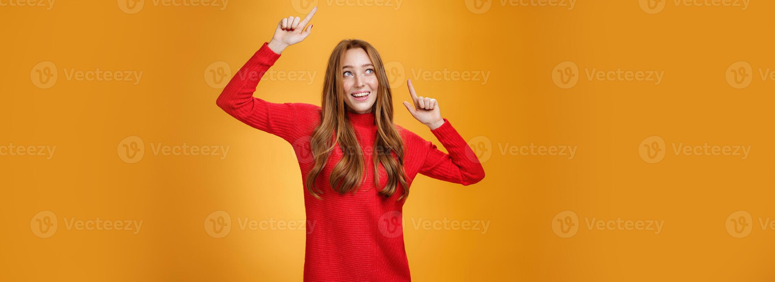 Charming and elegant attractive redhead female in red knitted dress raising hands up carefree as dancing having fun being pleased and happy gazing amused with broad grin at upper left corner photo