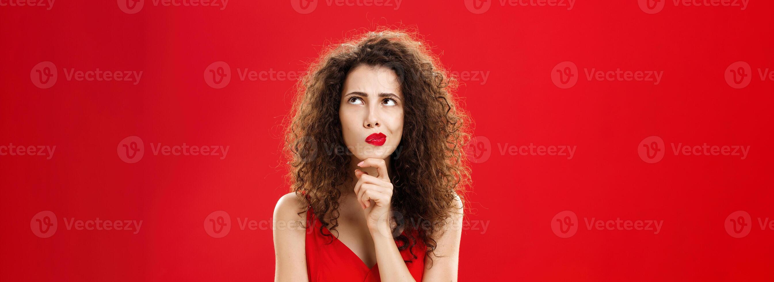 Smart and creative wife trying make up plan how steal money from bank account standing in thoughtful pose over red background in elegant dress smirking frowning looking at upper right corner concerned photo