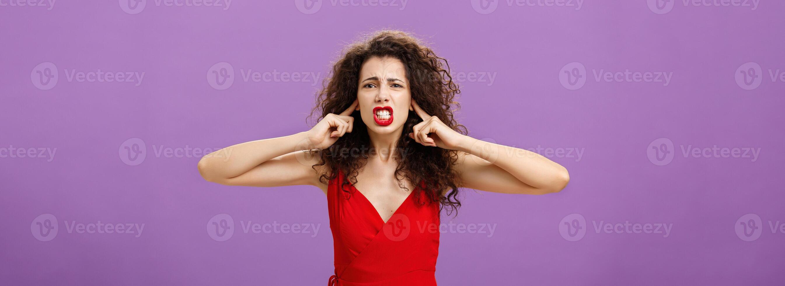 Woman fed up of hearing cursing words and arguments in her turn. Portrait of displeased insecure and offended female with curly hairstyle in red dress closing eyes and talking displeased and bothered photo