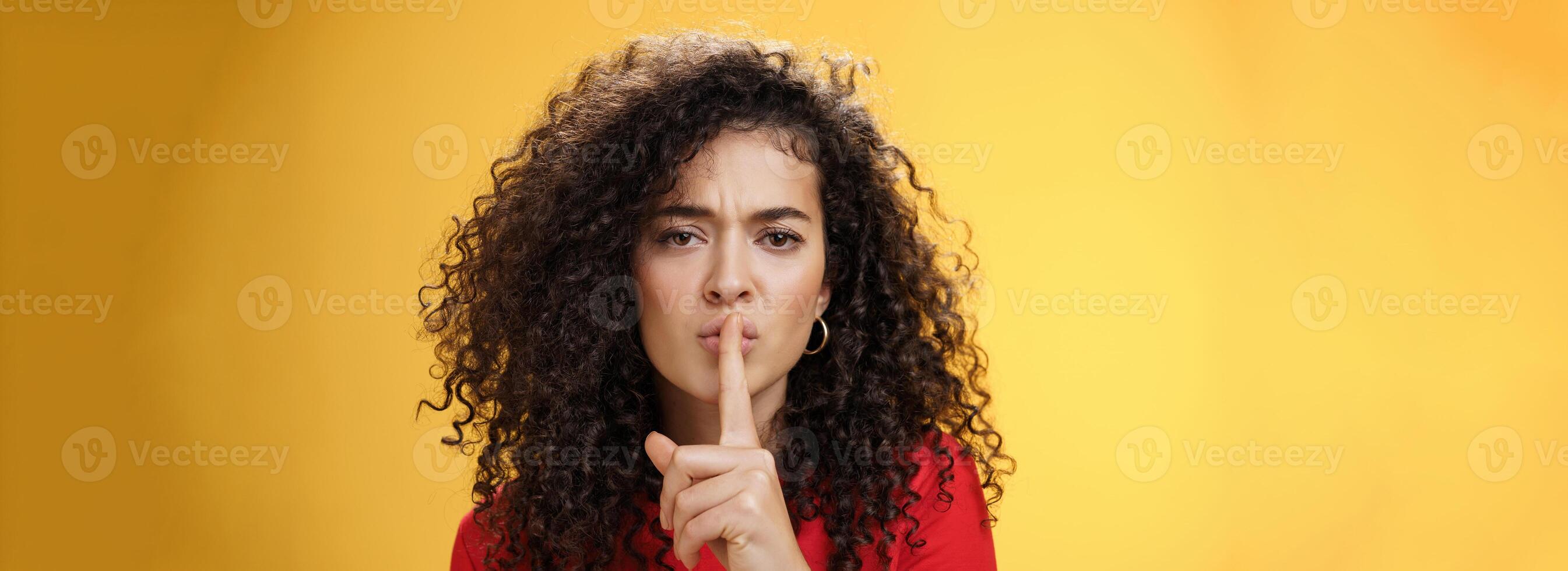 Shh not say word. Portrait of serious-looking sexy young woman with curly hair showing shush gesture with index finger over folded lips frowning asking keep secret promise keep silent over yellow wall photo