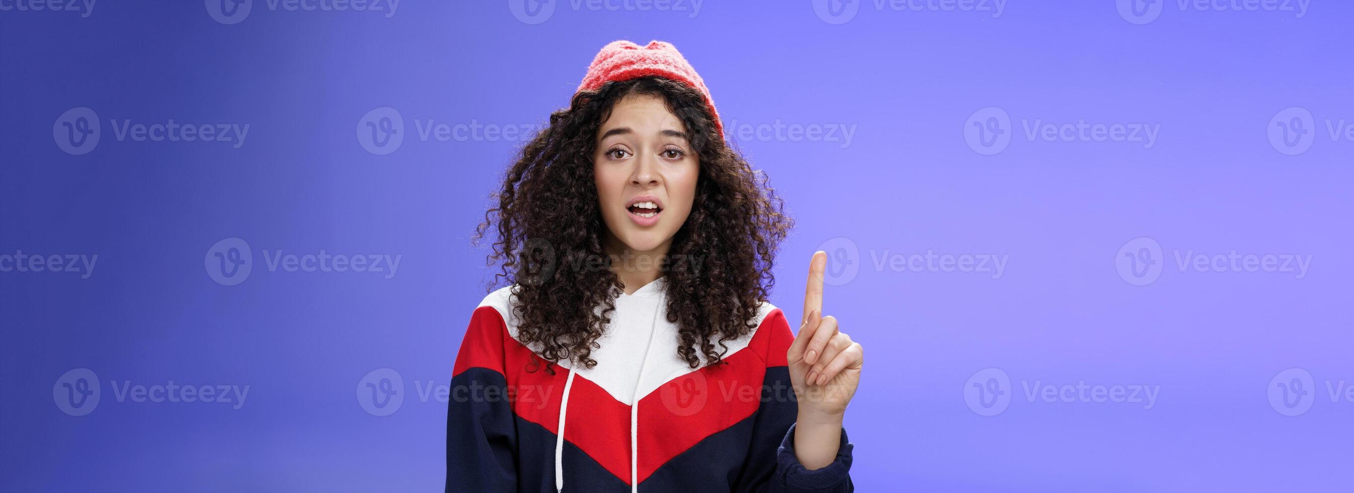 Rule number one do not mess with me babe. Portrait of swag and cool stylish young curly-haired woman in winter beanie showing index finger in prohibition or warning gesture over blue background photo