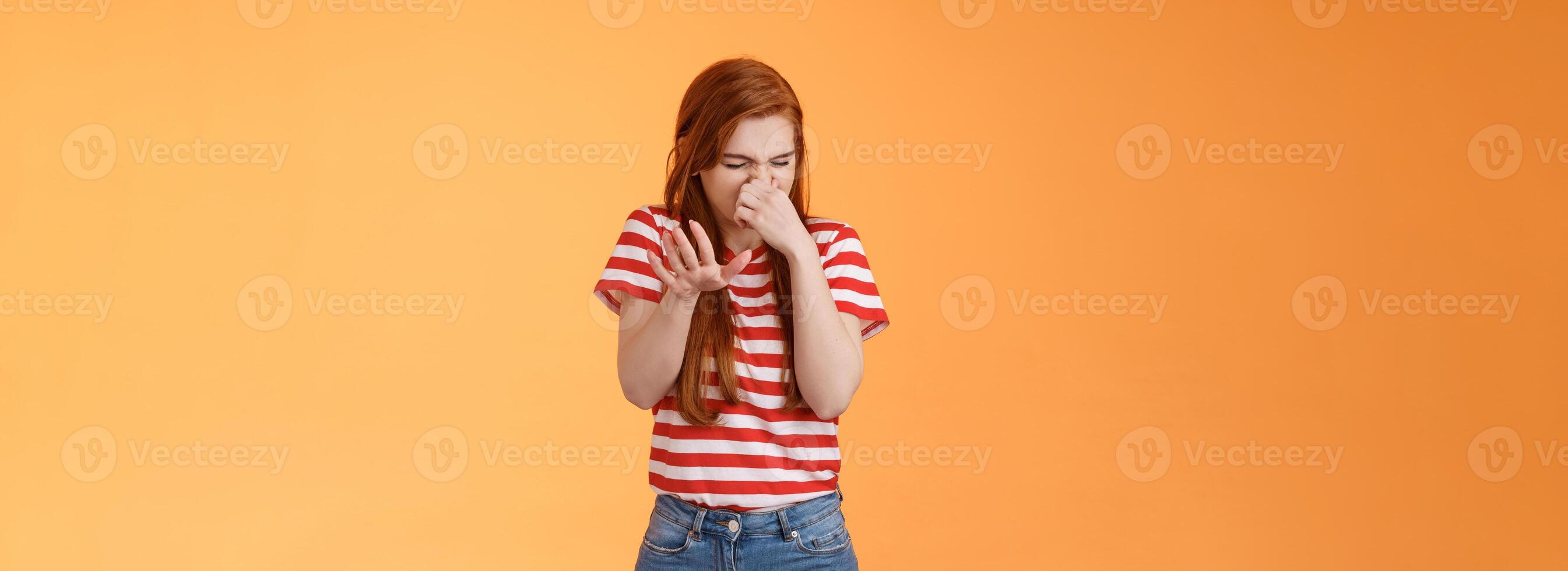 Gosh it stinks. Reluctant disgusted redhead woman raise hand block dislike gesture shut nose, cringe from aversion dislike, puke from reek bad smell, stand orange background disappointed photo