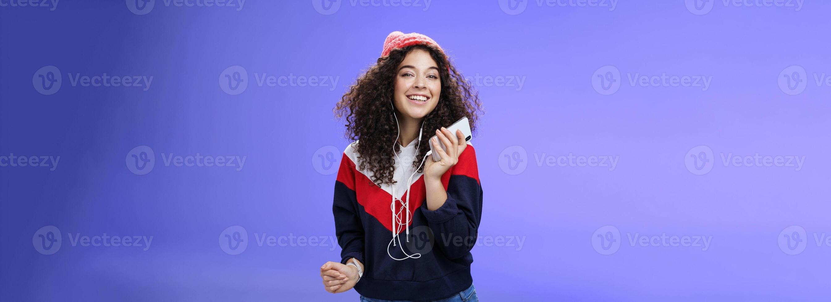 Waist-up shot of charming happy tender curly-haired female in beanie and sweatshirt listening music as dancing carefree, playing karaoke app or enjoying cool song in earphones over blue background photo