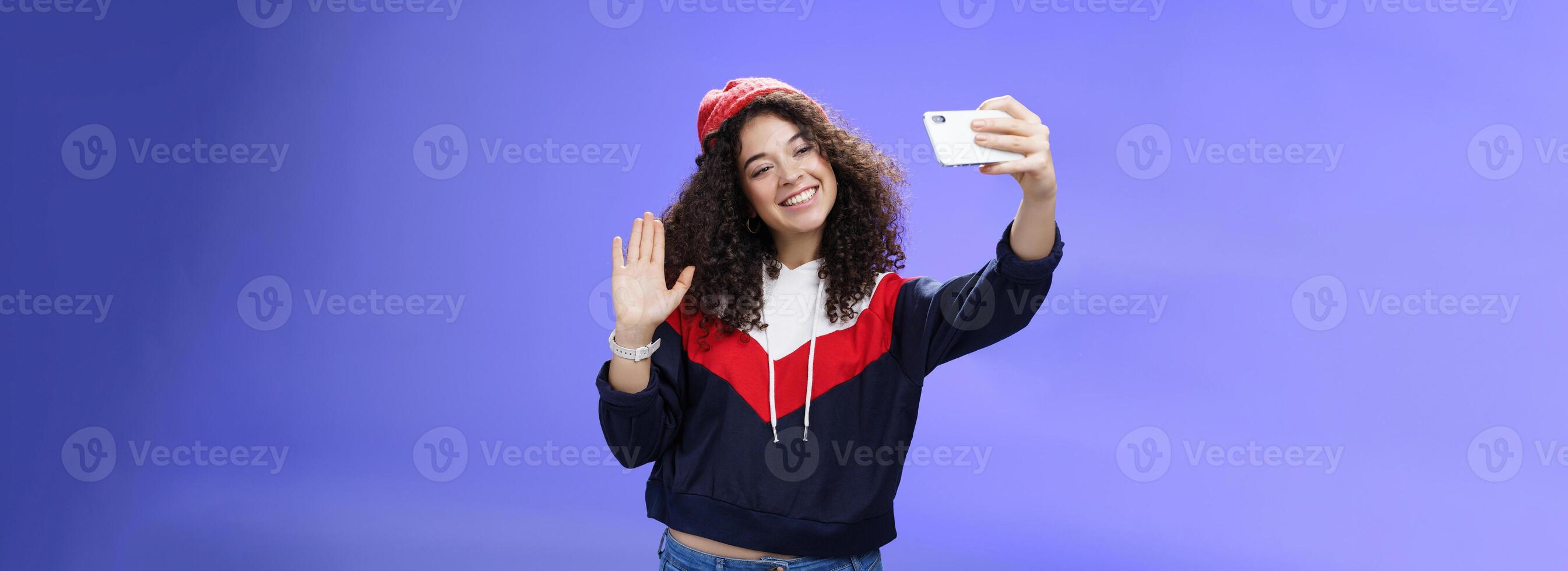Studio shto of stylish cute female vlogger with curly hair in winter red hat smiling and waving hello at smartphone camera as recording , making interesting content to post online over blue wall photo