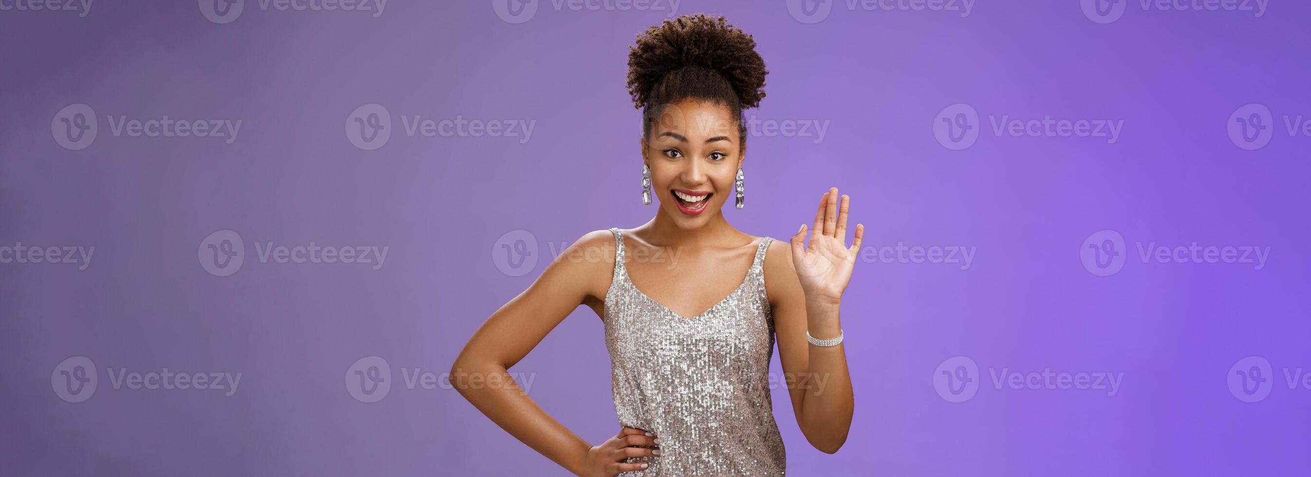 Friendly elegant african-american young 20s woman wearing elegant silver glittering dress hold hand waist waving hello greeting gesture say hi informally meet friend talking blue background photo