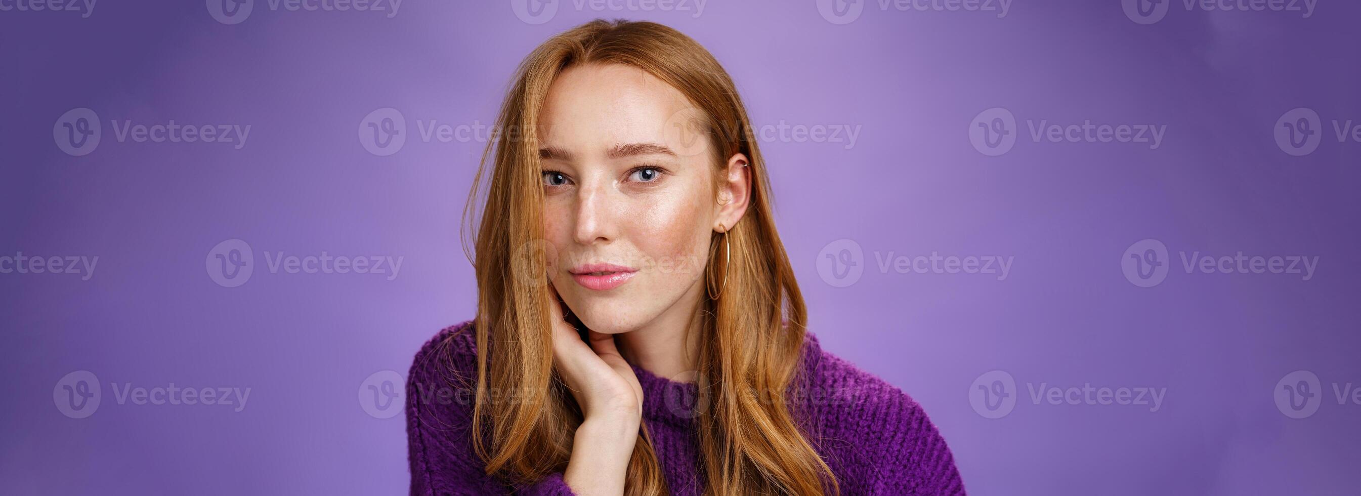 Sensual and romantic woman with red hair and freckles touching cheek gently as curls dropping on half face gazing charmed and flirty at camera, smiling being tender and coquettish over purple wall photo