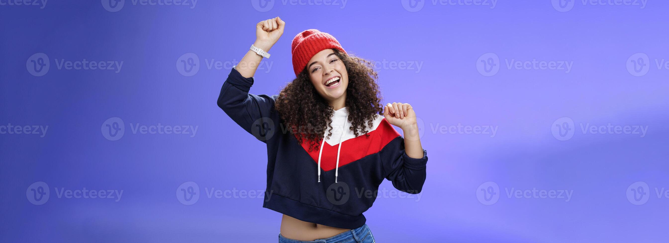 Hey come on dance with me. Friendly-looking bright and stylish cheerful woman with curly hairstyle wearing warm beanie raising hands up as enjoying great day, having fun outdoors over blue wall photo