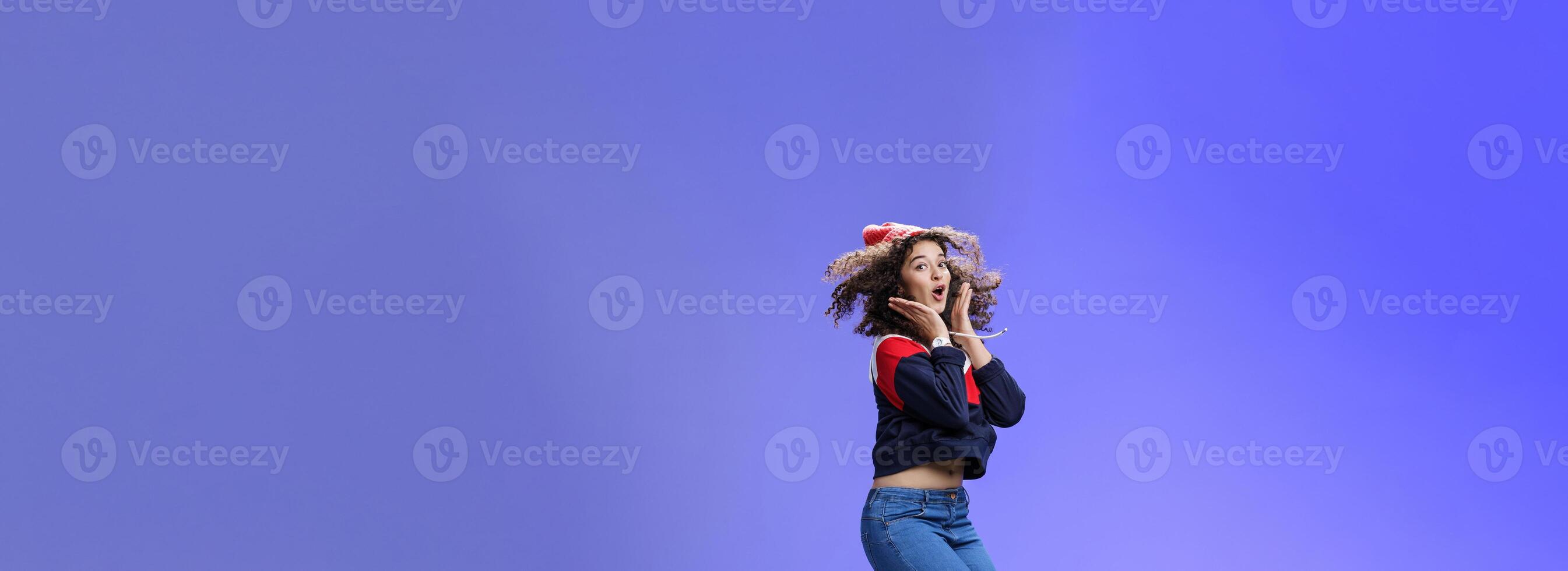 Surprised and amazed amused good-looking woman with curly hair in winter beanie and sweatshirt open mouth from joy and amazement jumping over blue background having fun feeling playful photo