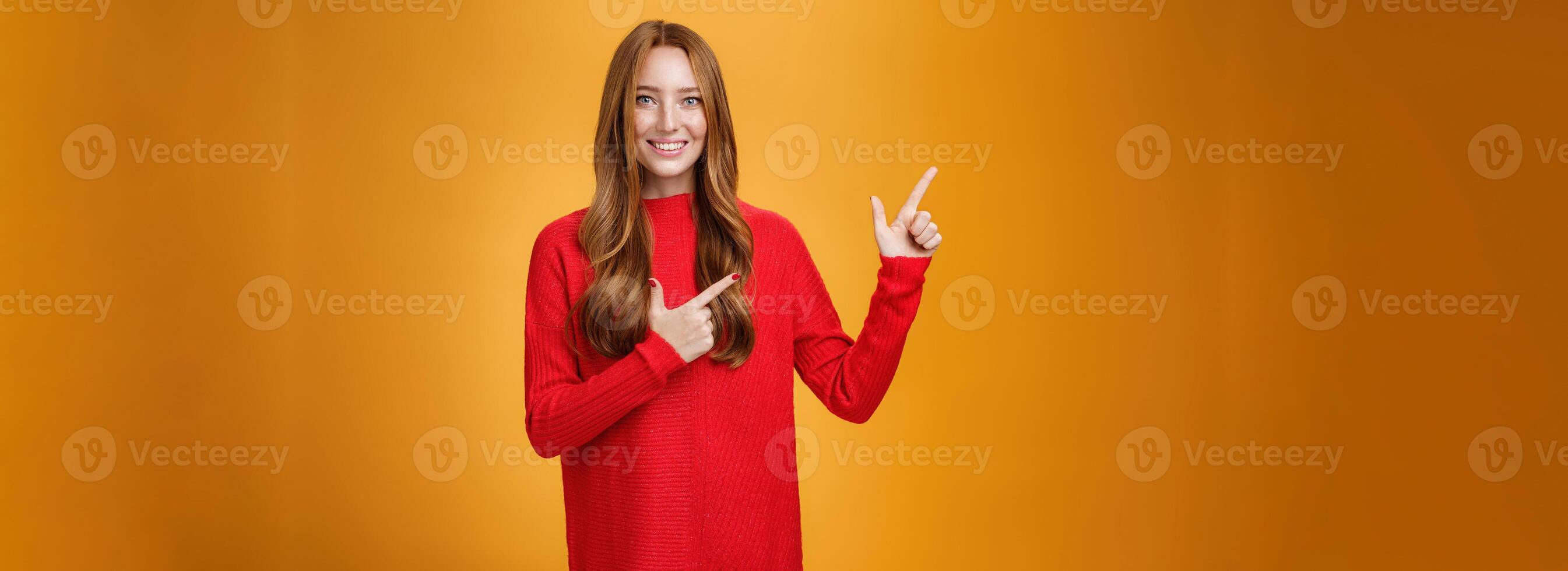 Assertive and helpful charming young female assitant in red knitted dress with ginger hair smiling friendly and pleasant as pointing at upper left corner, showing interesting thing over orange wall photo