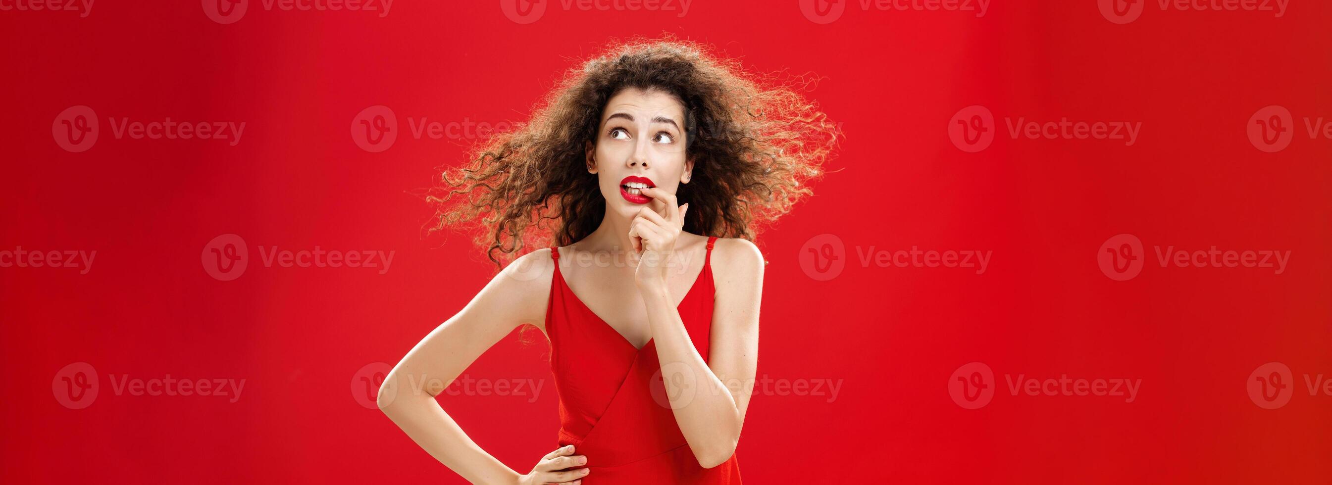 Waist-up shot of concerned and worried insecure cute female with curly hairstyle in red evening dress biting finger looking troubled or questioned at upper right corner with hand on waist photo