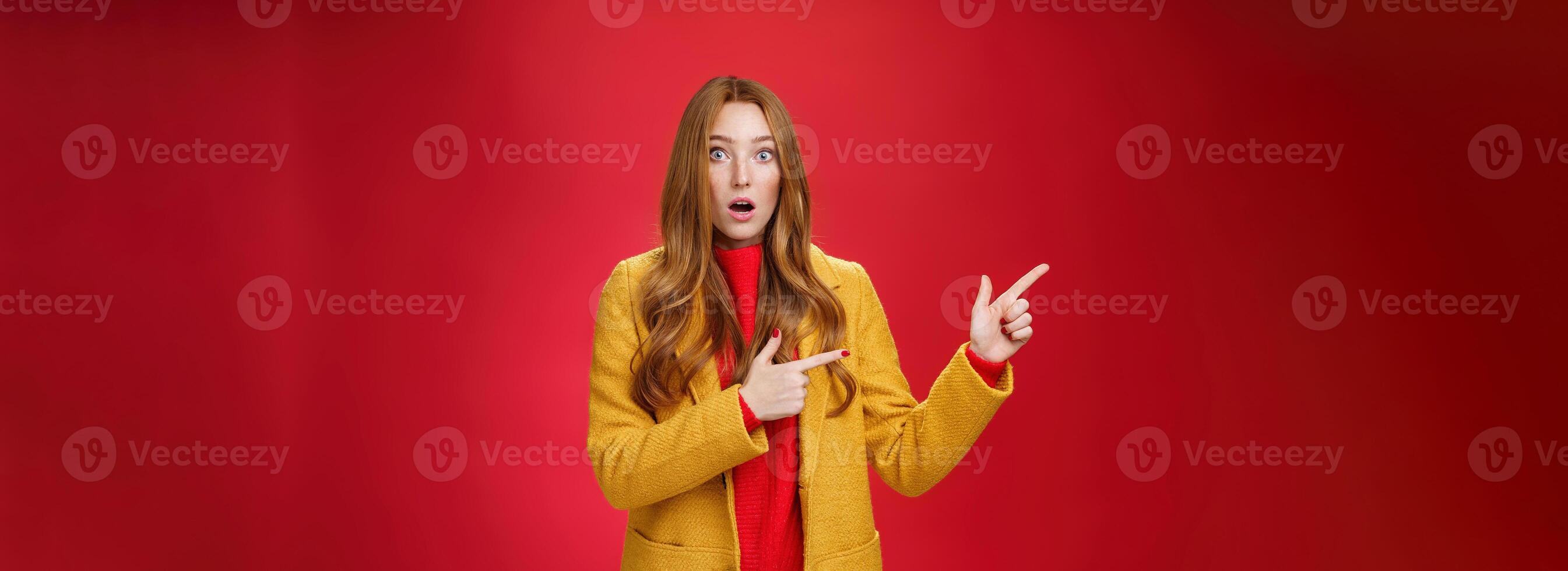 Stunned speechless and impressed attractive redhead woman with freckles in yellow coat drop jaw amazed as popping eyes at camera questioned, pointing left astonished over red background photo