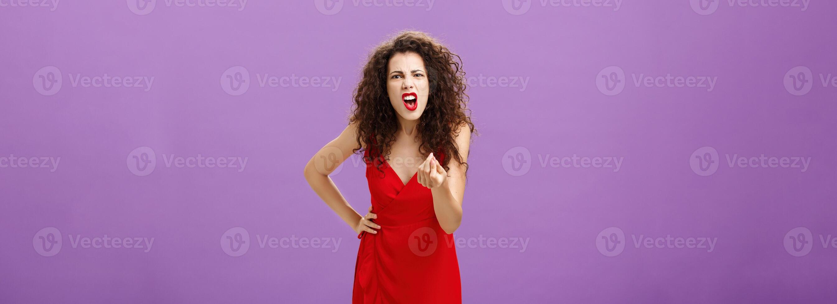 Woman in fury fighting with husband over money making italian gesture with fingers shouting and yelling from fury and anger standing displeased and pissed over purple wall in red elegant dress photo