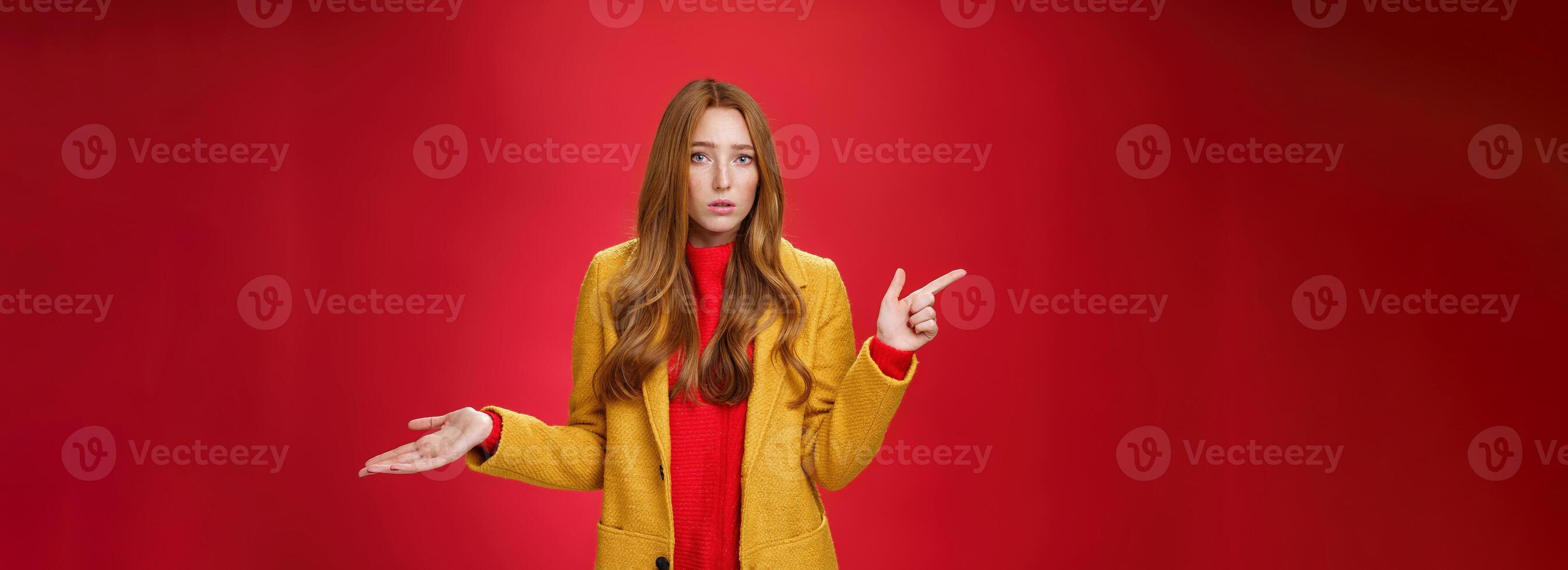 Frustrated and confused young cute female with ginger hair in yellow coat shrugging as pointing questioned left waiting explanation as asking question clueless and puzzled over red background photo