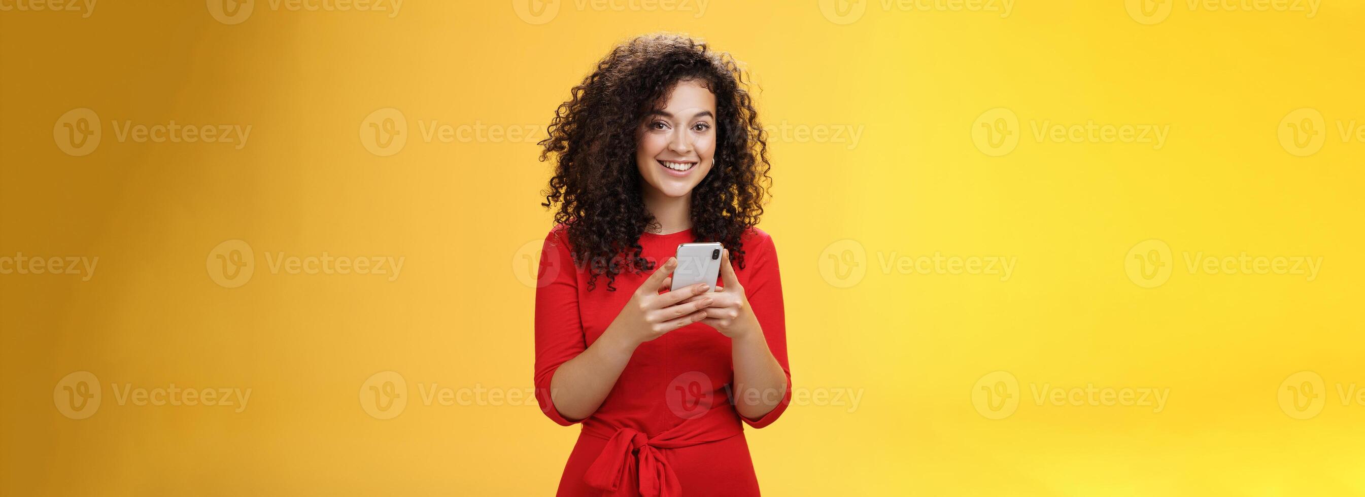 Girl messaging friend to tell all hot rumors holding mobile phone in hands smiling broadly and excited at camera as using smartphone browsing in net or edditing in app over yellow background photo