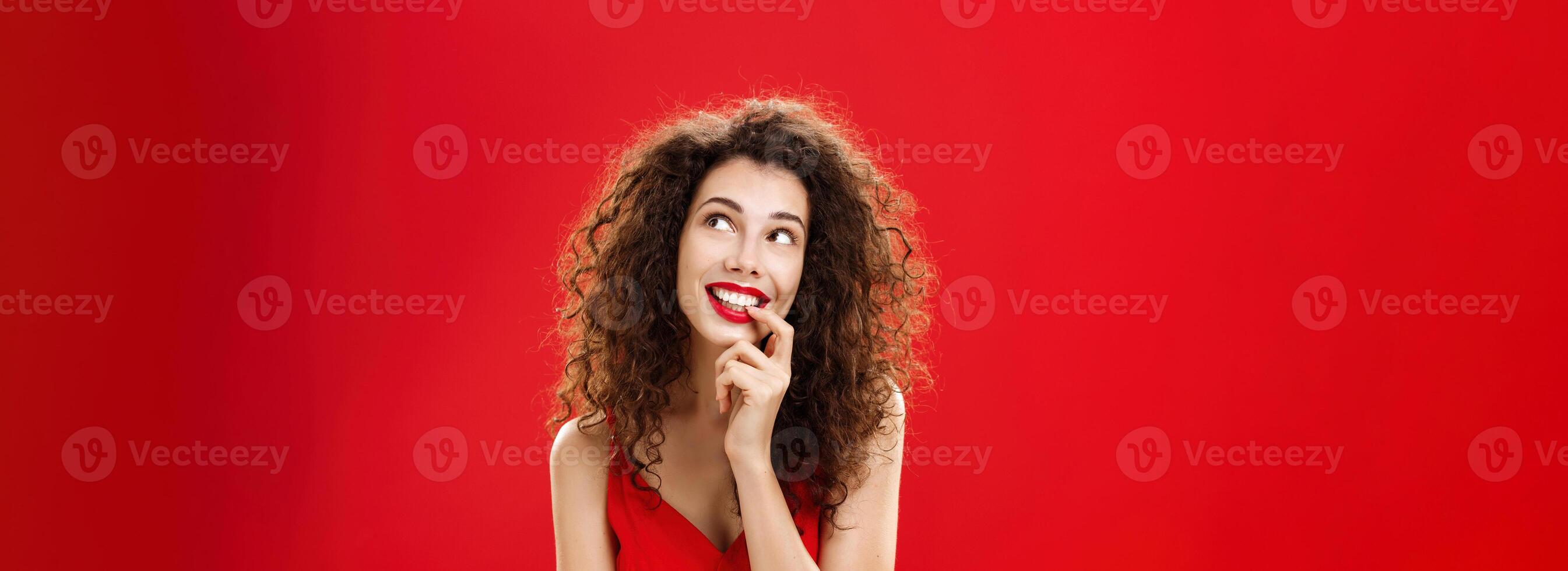 Cute charismatic and tender dreamy woman with curly hairstyle in red dress biting finger and smiling with desire in eyes looking at upper right corner daydreaming picturing object she wants photo