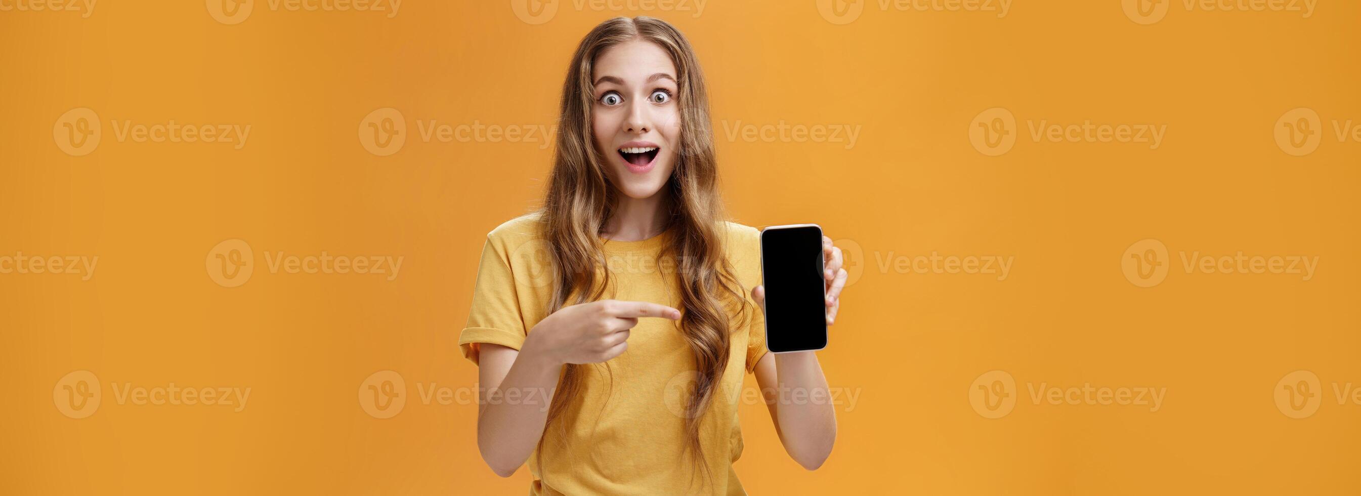 Girl surprised with awesome smartphone features. Portrait of delighted and amazed cute european slender woman with wavy natural hairstyle showing cellphone pointing at gadget screen at app photo