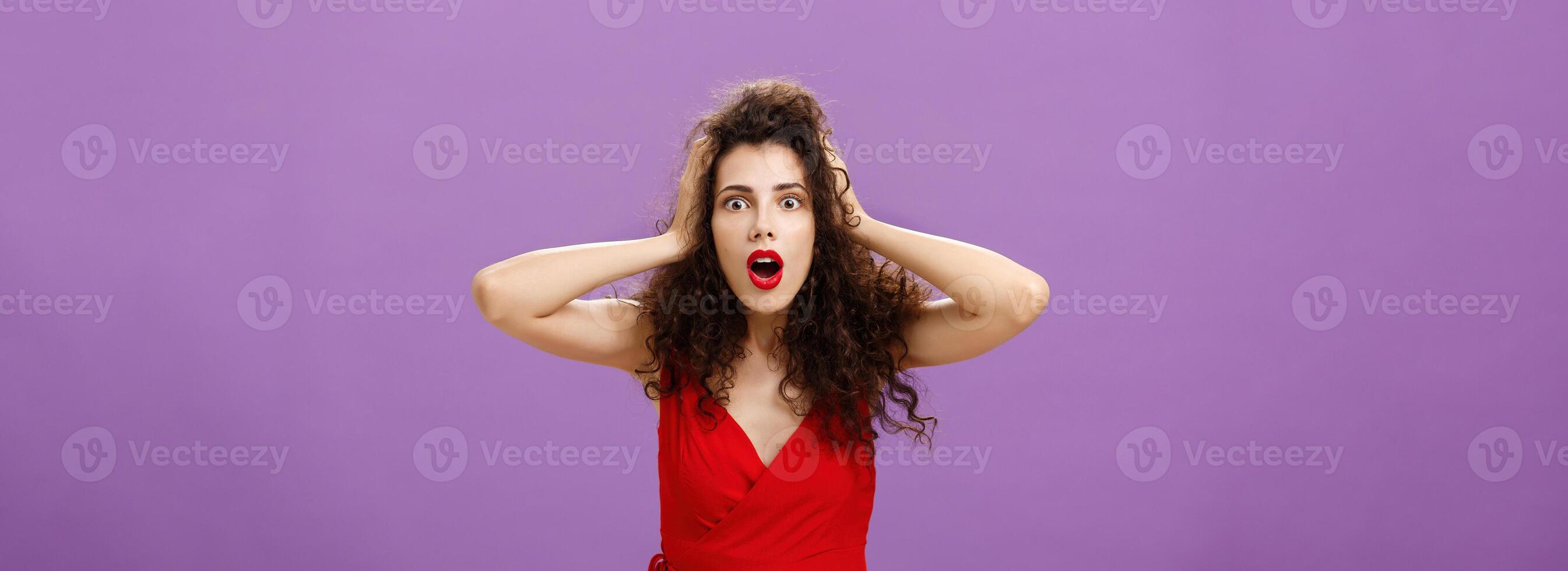 You do not say. Portrait of shocked and impressed speechless woman with curly haircut in luxurious evening dress opening mouth from amazement and shock holding hands on head stunned over puple wall photo