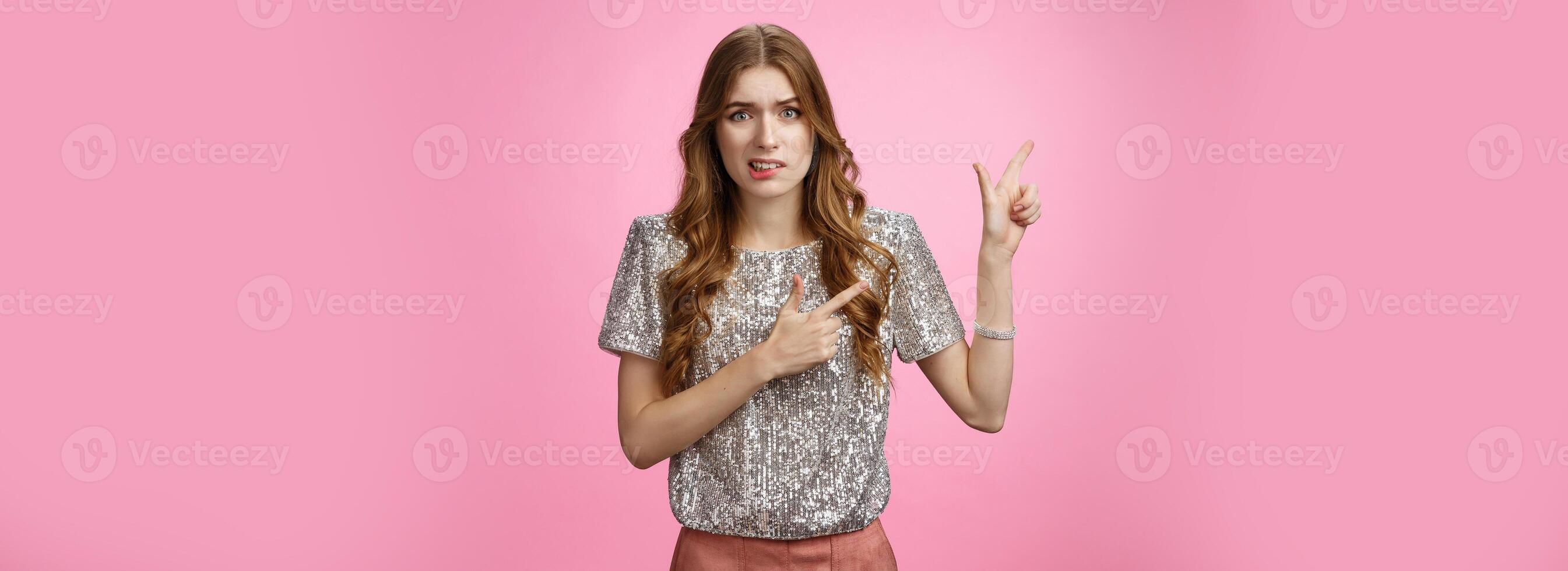 Dispelased insecure awkward young glamour rich girl dress-up party disappointed dislike place cringing aversion disgust pointing left questioned troubled, concerned standing pink background photo