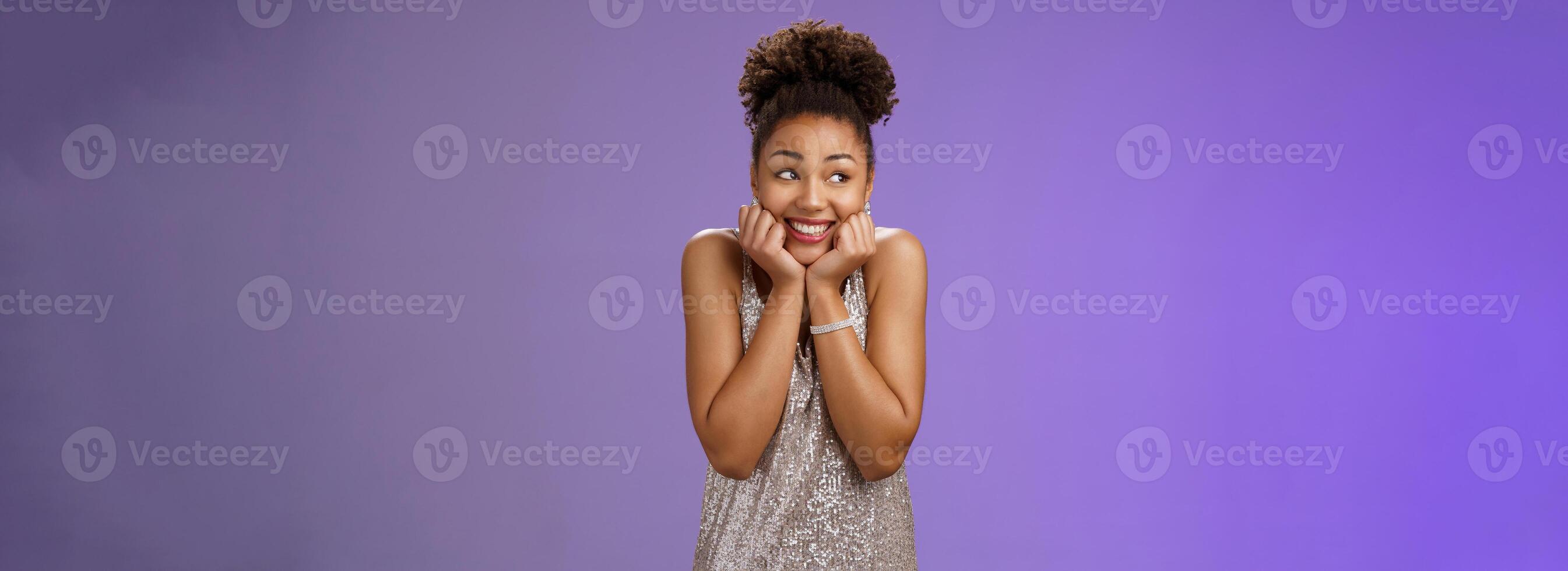 Amused silly cute black girl in silver dress anticipating exciting party dreamy gaze up touch cheeks smiling broadly optimistic waiting dream come true standing pleased touched blue background photo