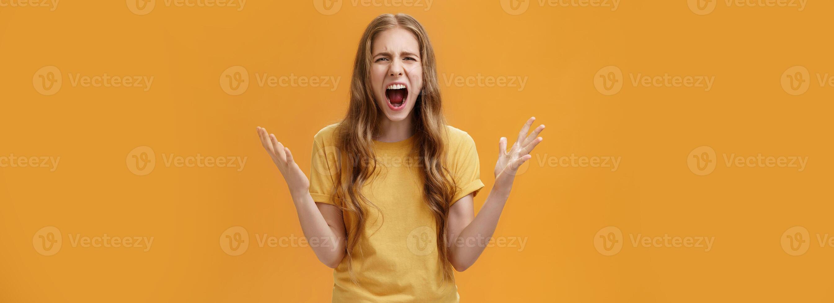 Studio shot of young woman during argument losing temper standing pressured and pissed yelling out loud from hate and anger gesturing with raised palms making furious face posing against orange wall photo