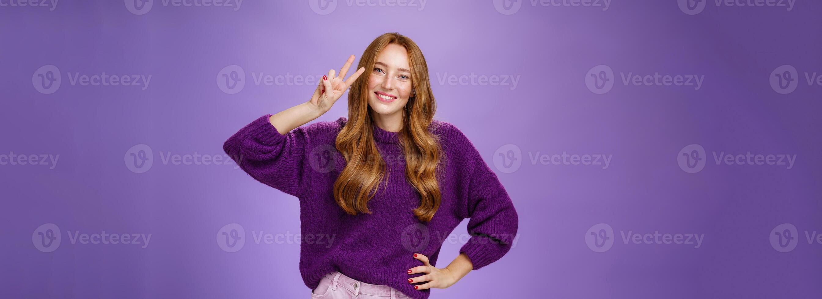 Attractive energized redhead woman in purple sweater showing victory gesture near eye and smiling broadly tilting head satisfied and joyful, holding hand on waist posing over violet background photo