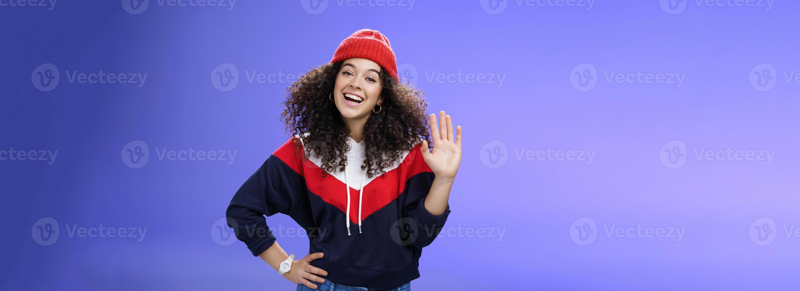 Hey nice to meet you. Charming female ski couch in cute red beanie with curly hair waving hello with palm and smiling broadly greeting newbies as teaching winter sports over blue background photo
