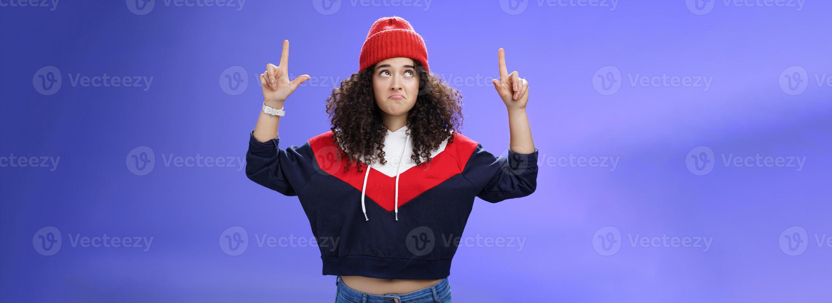 Girl feeling disappointed and gloomy as looking at bad weather making sad smile, pursing lips down raising hands staring up upset and unsatisfied standing displeased against blue background photo