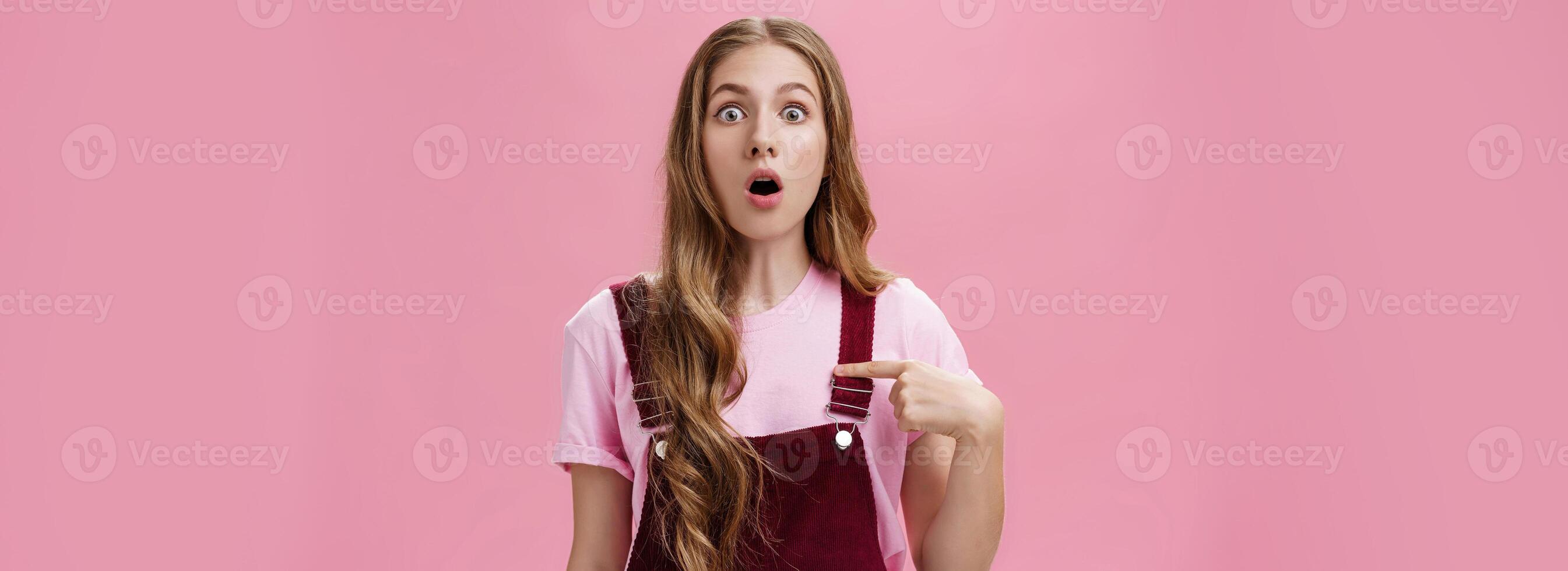 Shocked uncertain and confused charming young woman with tattoos and long natural wavy hair dropping jaw gasping and popping eyes from surprise pointing at herself with unsure expression photo