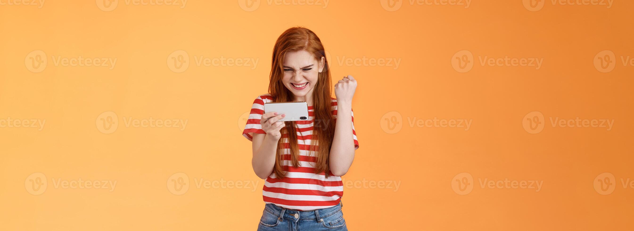 Pleased lucky redhead girl gamer redhead, fist pump satisfied, say yes excited stare display, hold smartphone horizontal, pass hard level playing awesome game use application, achieve victory photo