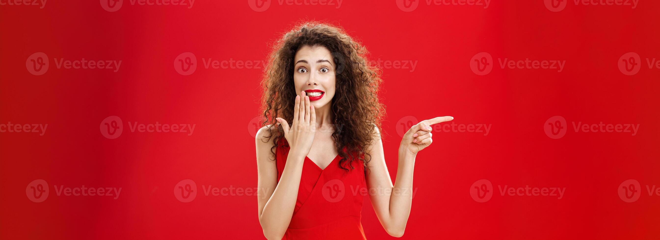 Troubled cute and tender silly woman with curly hairstyle in red stylish evening dress saying oops smiling with guilty look covering mouth with palm while pointing left over studio background photo