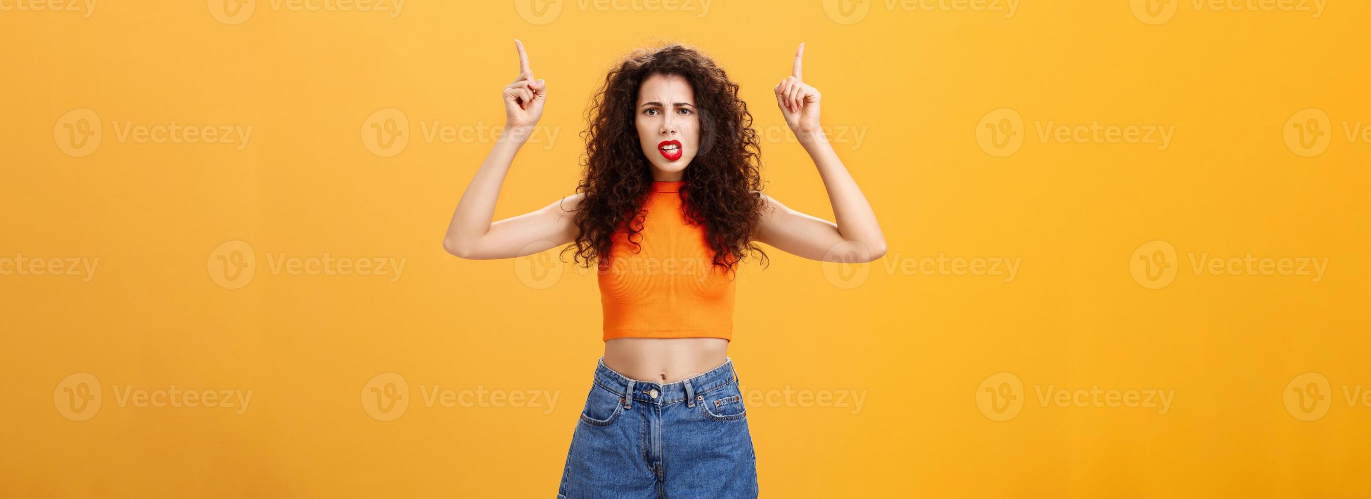 Indoor shot of displeased female client with curly stylish hairstyle red lipstick in cool cropped top pointing up demanding explanation with confused and dissatisfied expression over orange background photo