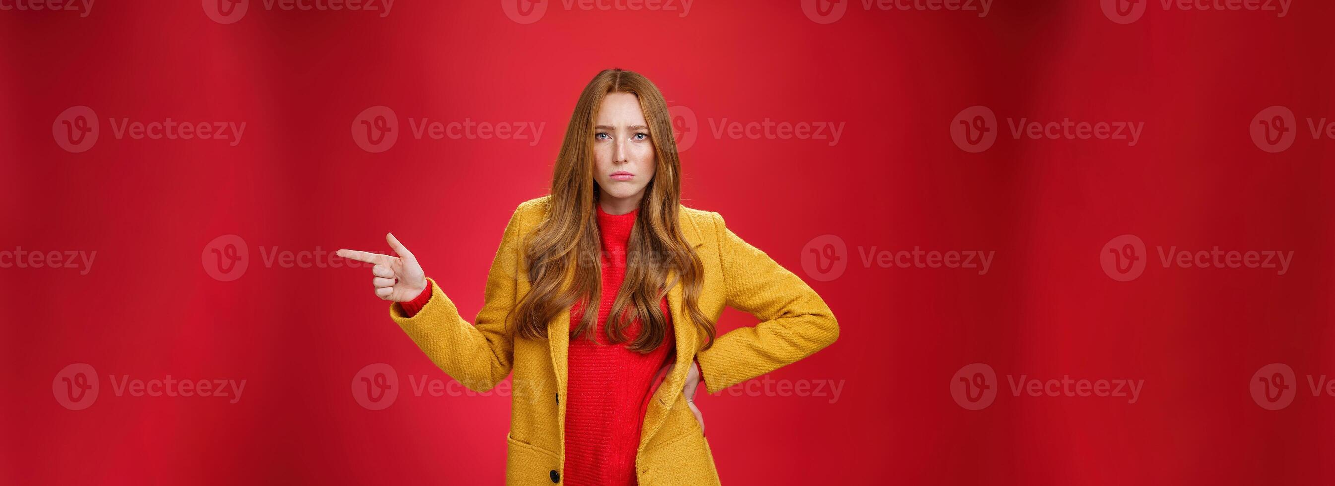 Explain yourself now. Portrait of upset gloomy and sad cute redhead girl waiting explanations asking question, pouting offended and pointing right with finger looking unhappy at camera over red wall photo
