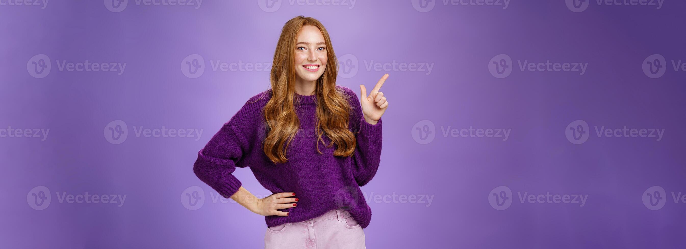 Hey click here. Portrait of friendly-looking charming and cute redhead female in purple sweater pointing at upper left corner with index finger and smiling, showing copy space, recommending, offering photo