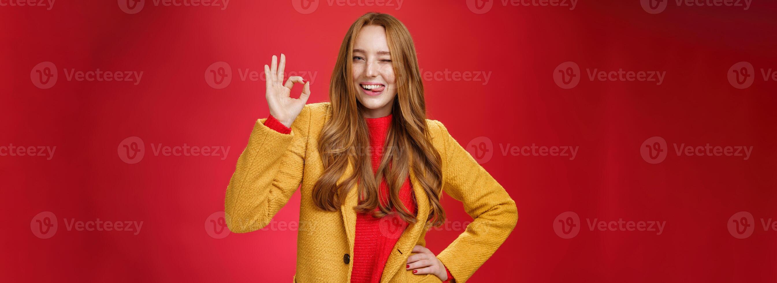 Okay got it. Portrait of carefree happy and confident good-looking charismatic ginger woman with freckles winking and showing tongue joyfully showing ok gesture, liking excellent work over red wall photo
