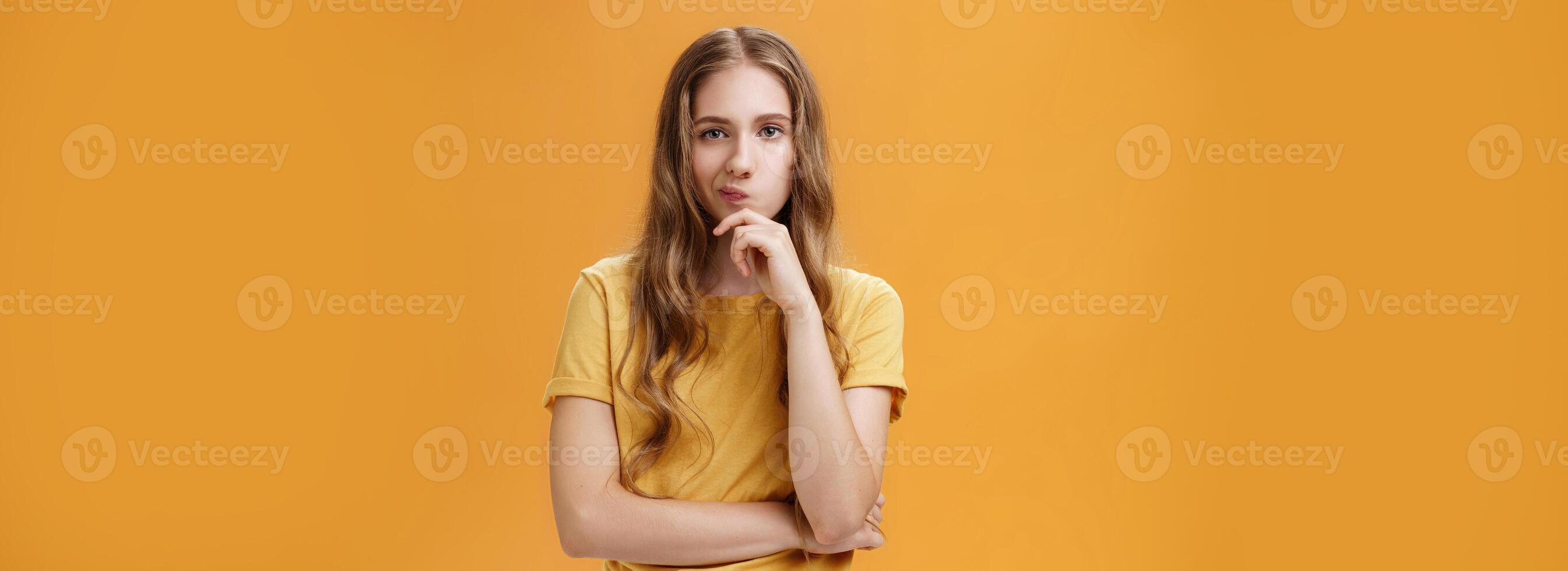 Hmm choices difficult. Portrait of focus serious-looking creative young female with wavy hair smirking holding hand on chin in thoughtful pose, making up decision, thinking over orange background photo