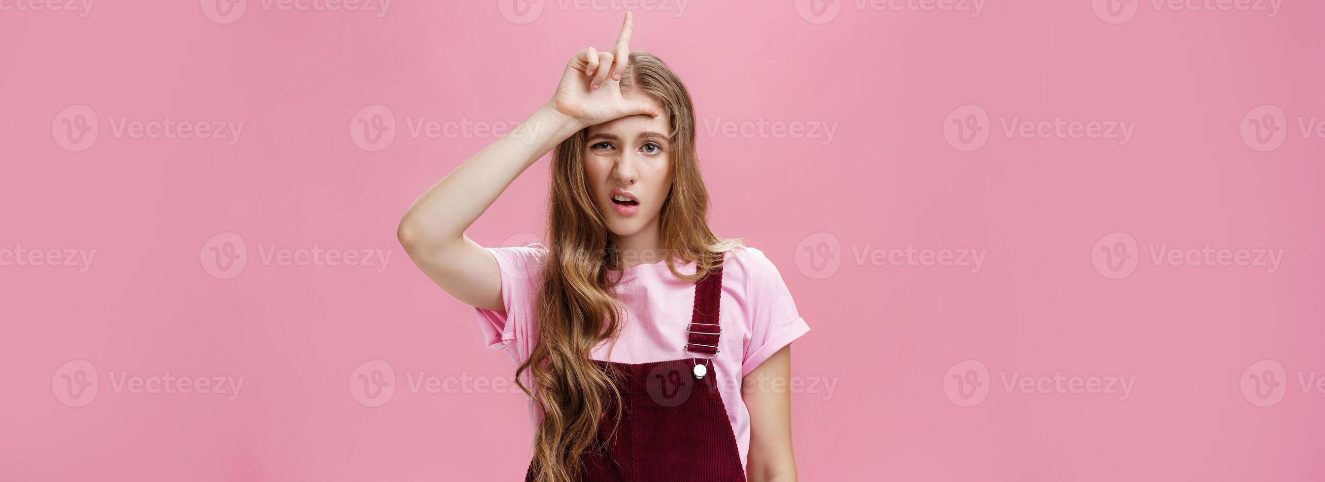 Waist-up shot of arrogant young girl with high ego grimacing snobbish showing letter L on forehead mocking over guy who lose calling person loser with scorn and disdain over pink background photo