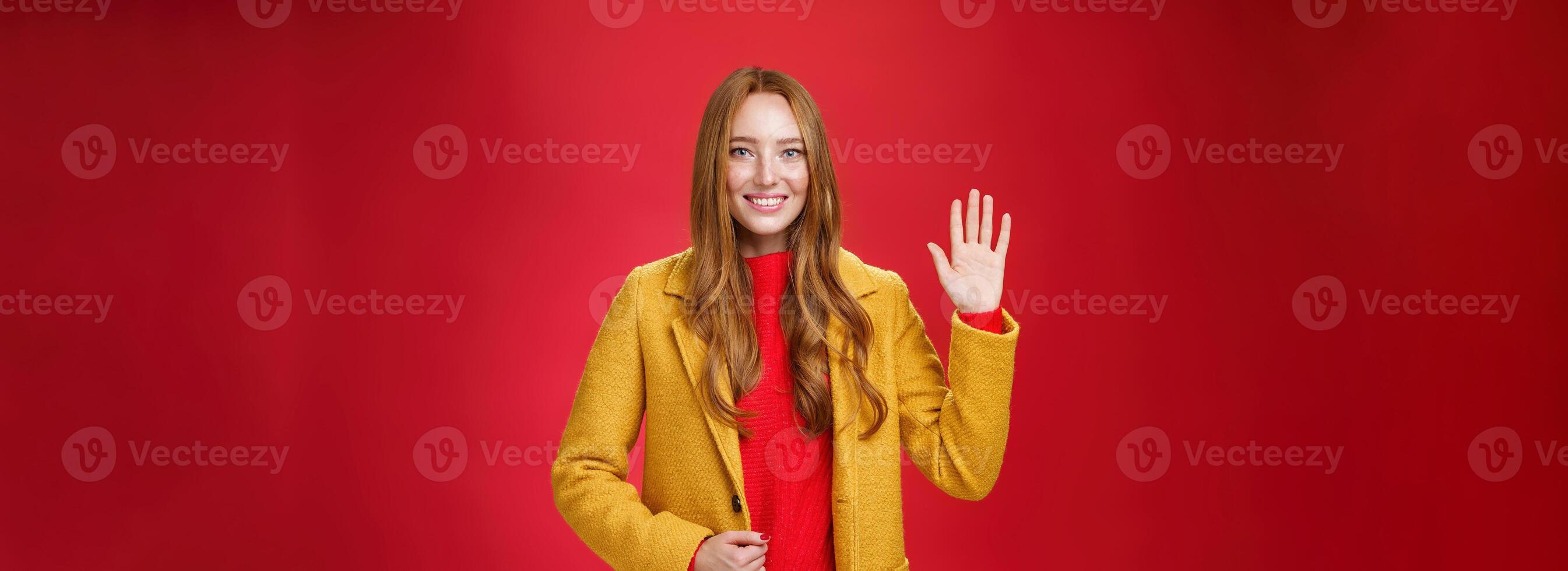 Hello nice meet you pal. Friendly-looking feminine and stylish young cute redhead female in yellow warm autumn coat waving raised hand in greeting and hi gesture smiling broadly over red wall photo