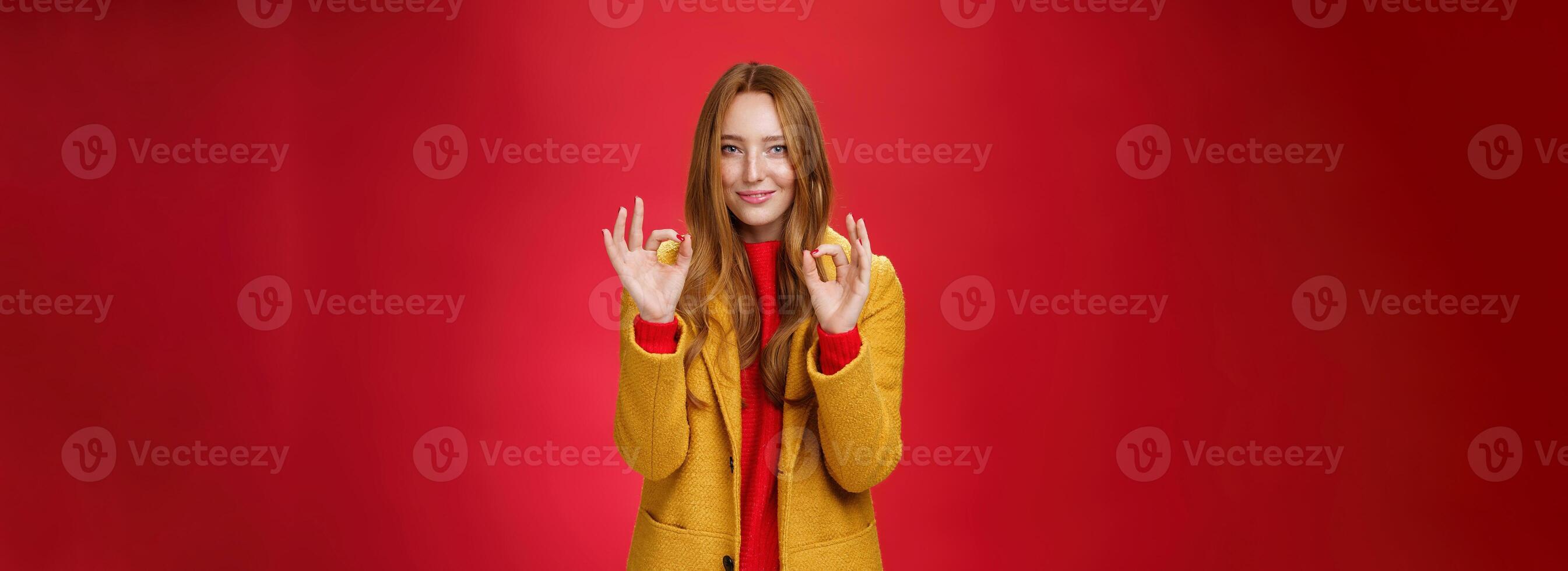 Indoor shot of pleased bright and happy young woman feeling okay and perfect in warm comfortable yellow coat picking right close for cold weather showing ok gesture and smiling delighted over red wall photo