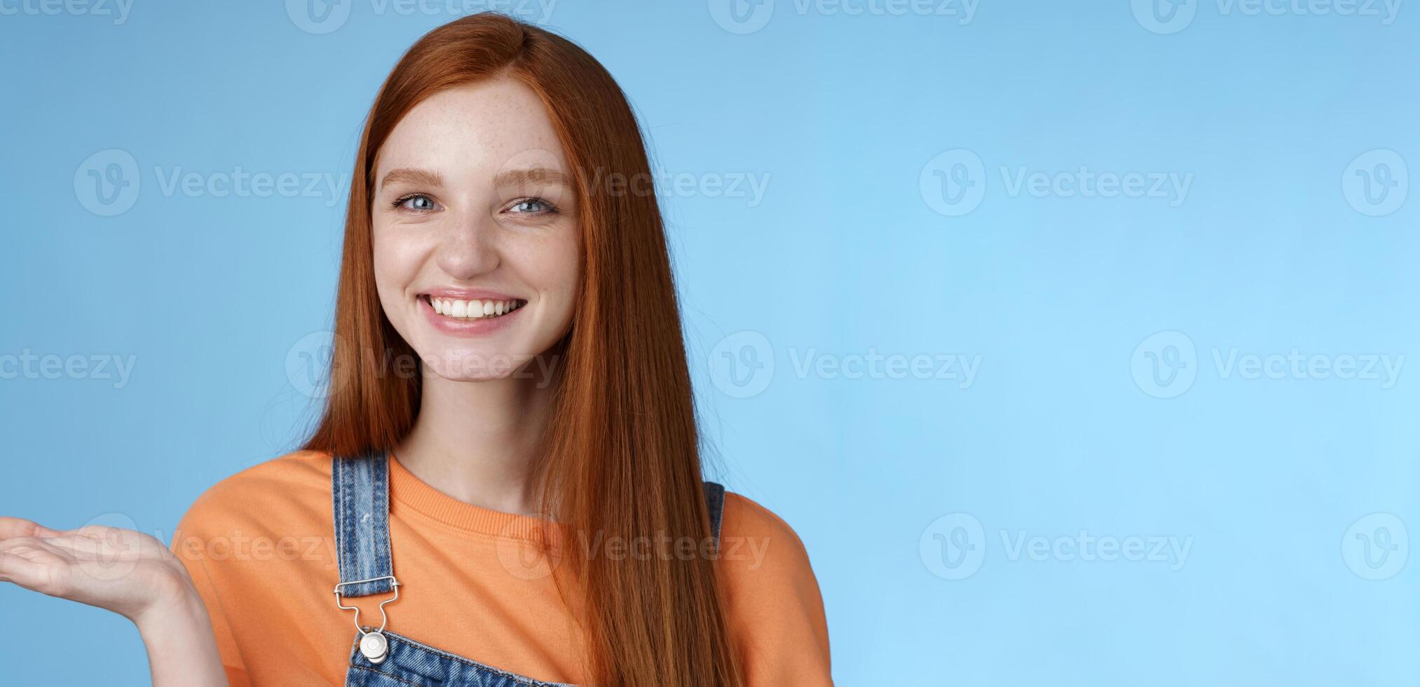 Proud good-looking confident redhead girl present awesome product hold object palm raise hand blank blue copy space smiling delighted recommend cool link, standing studio background helpful photo