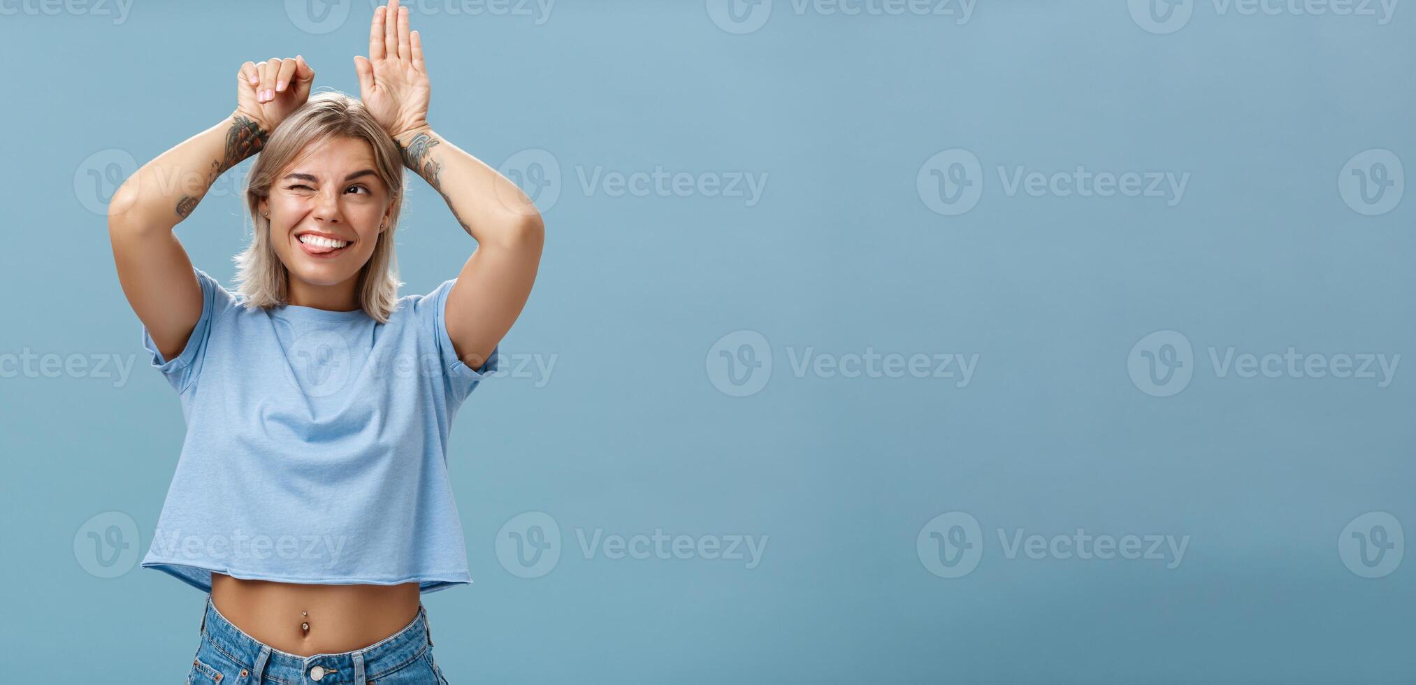 Studio shot of entertained carefree and emotive happy charming woman with tattoos on arms acting like bunny with palms on head winking joyfully smiling and sticking out tongue over blue wall photo