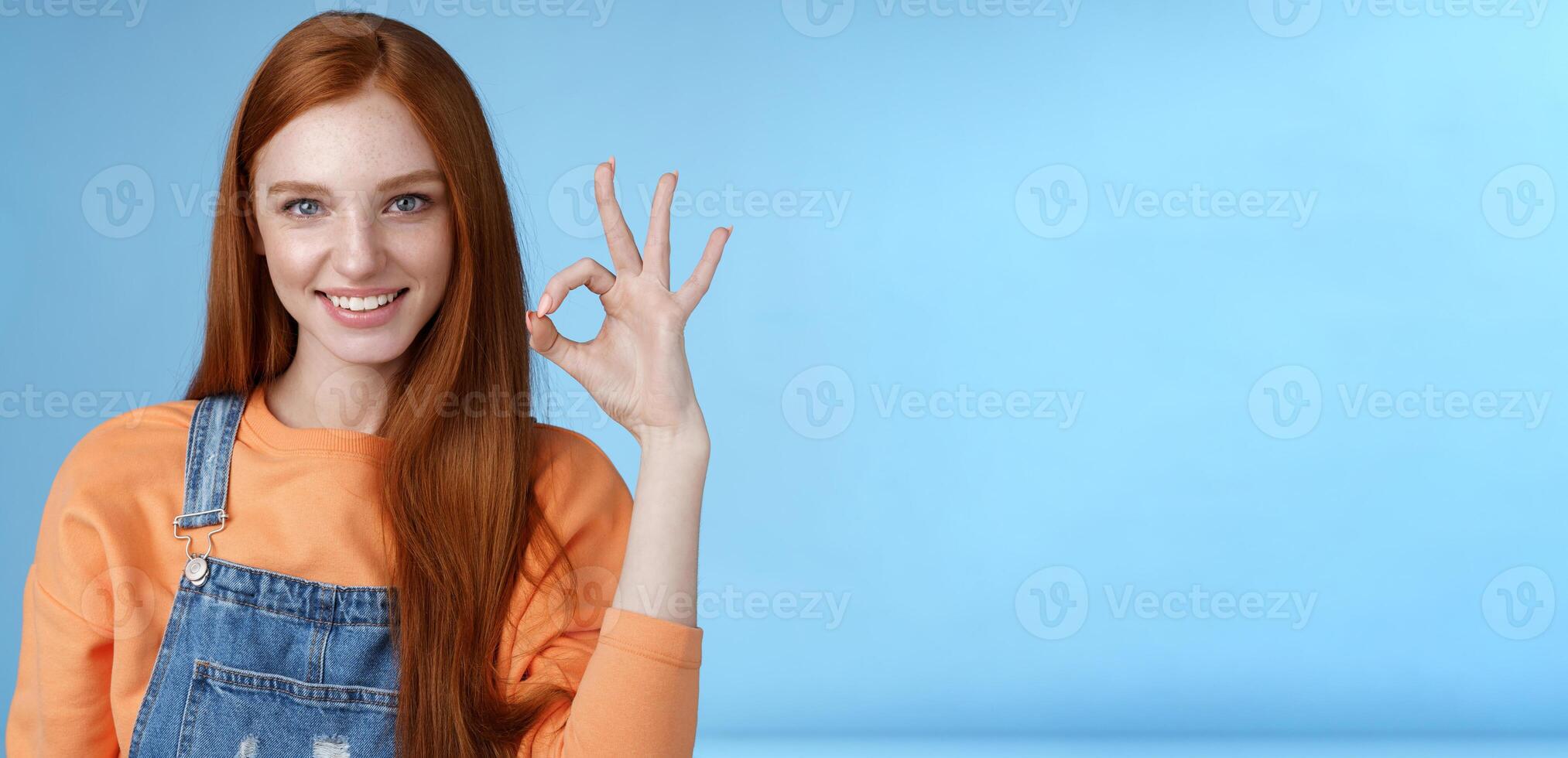 Sassy confident redhead girl got under control show okay ok excellent gesture cheeky look camera grinning assertive assuring everything awesome give permission approve good product, blue background photo