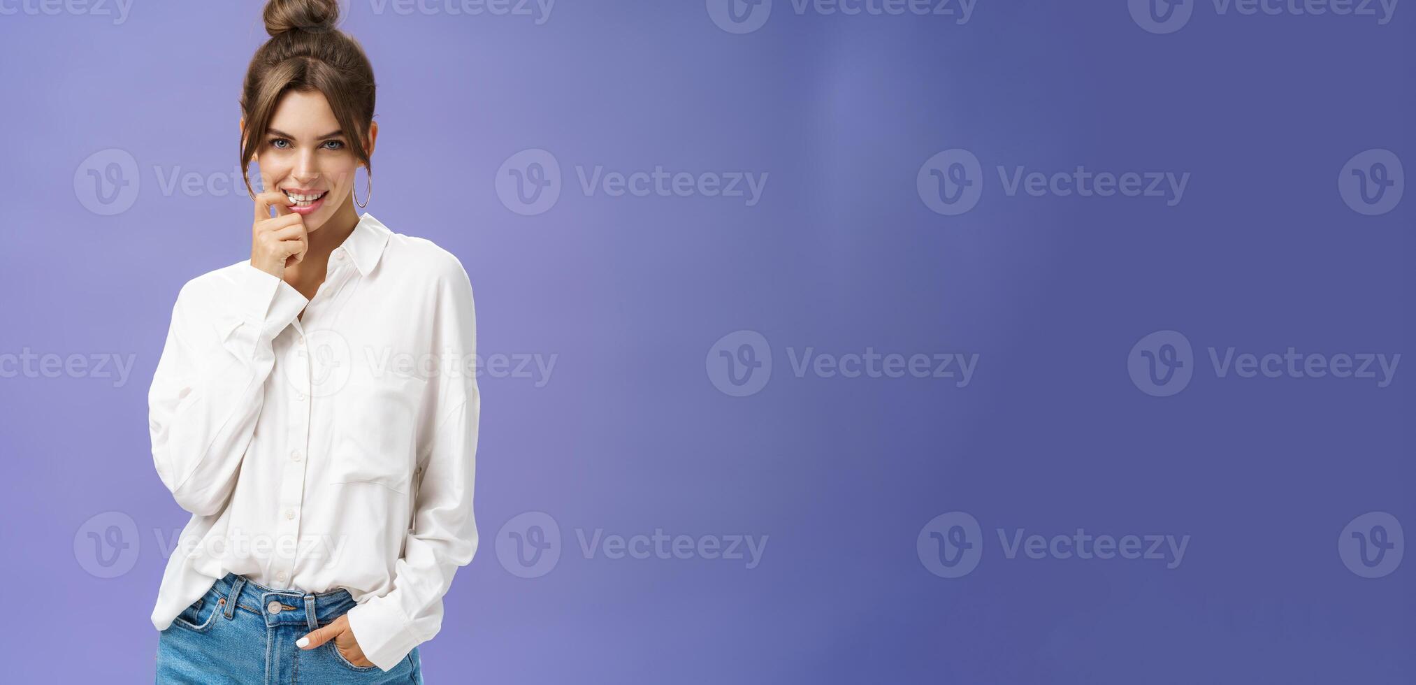 Sensual and flirty self-assured attractive woman in stylish white blouse with good self-esteem holding hand in pocket biting finger showing seducing skills looking daring at camera over purple wall photo