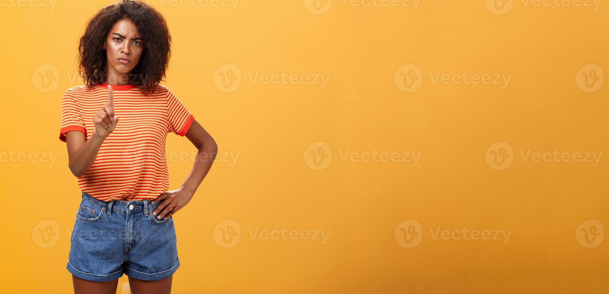 You better not. Dissatisfied bossy serious-looking african american curly-haired female head manager in t-shirt frowning shaking index finger in forbid or stop gesture scolding dog over orange wall photo