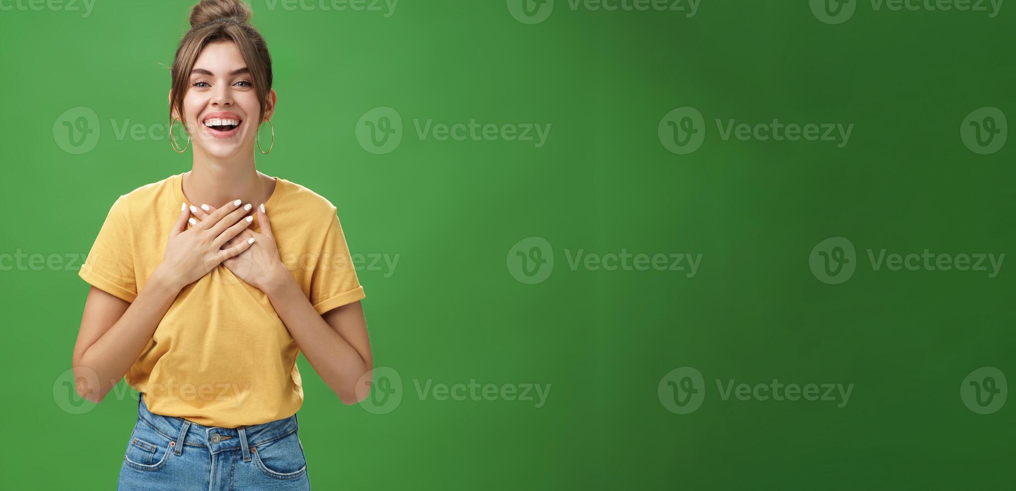 Delighted charming woman with gapped teeth in cozy outfit holding palms on chest in grateful or thankful pose smiling broadly receiving warm congratulations standing satisfied, happy over green wall photo