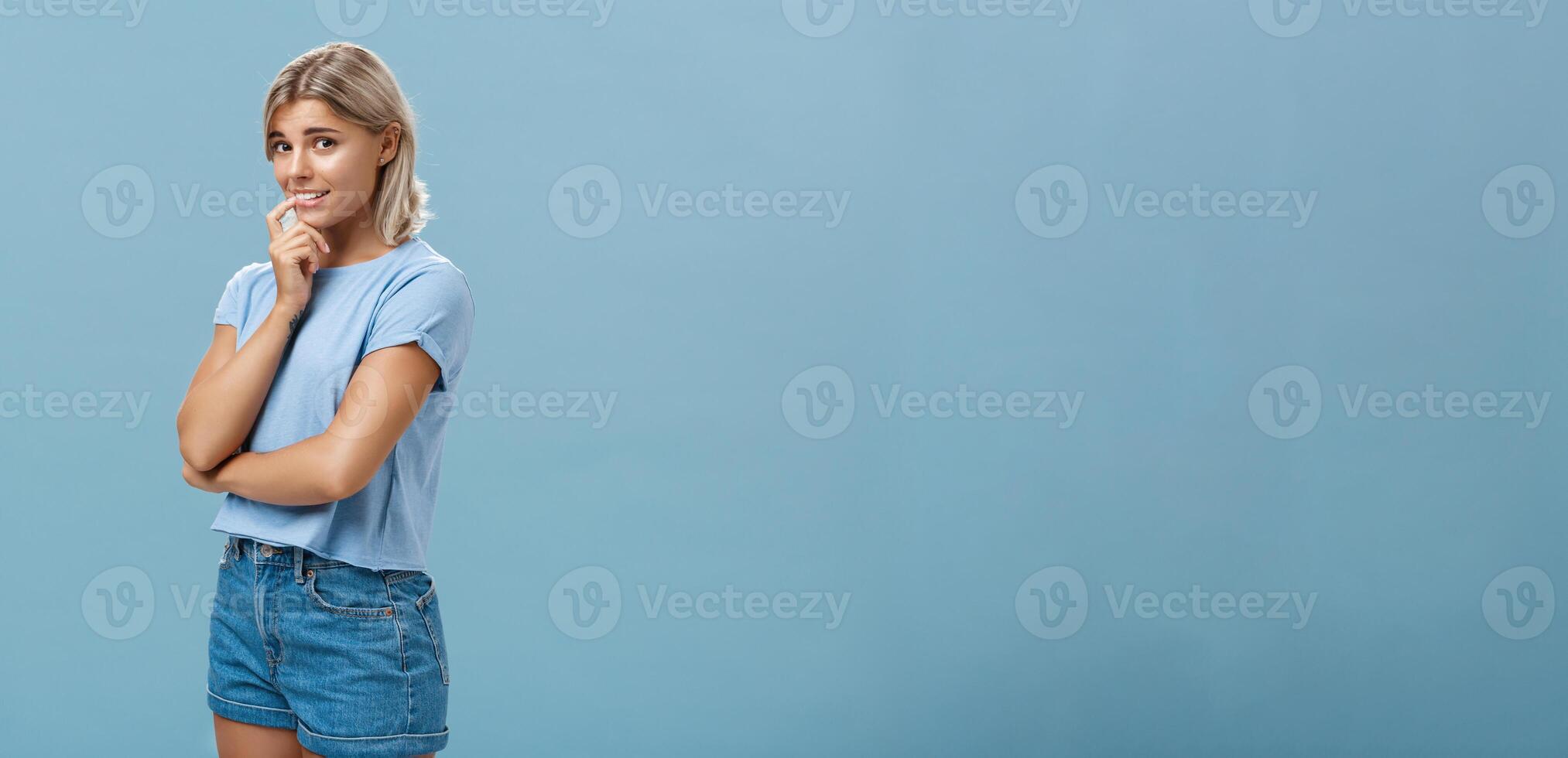 Studio shot of unconfident silly and insecure cute feminine girl with blond hair standing half-turned over blue background holding finger on lip while being shy ask question smiling hesitating photo