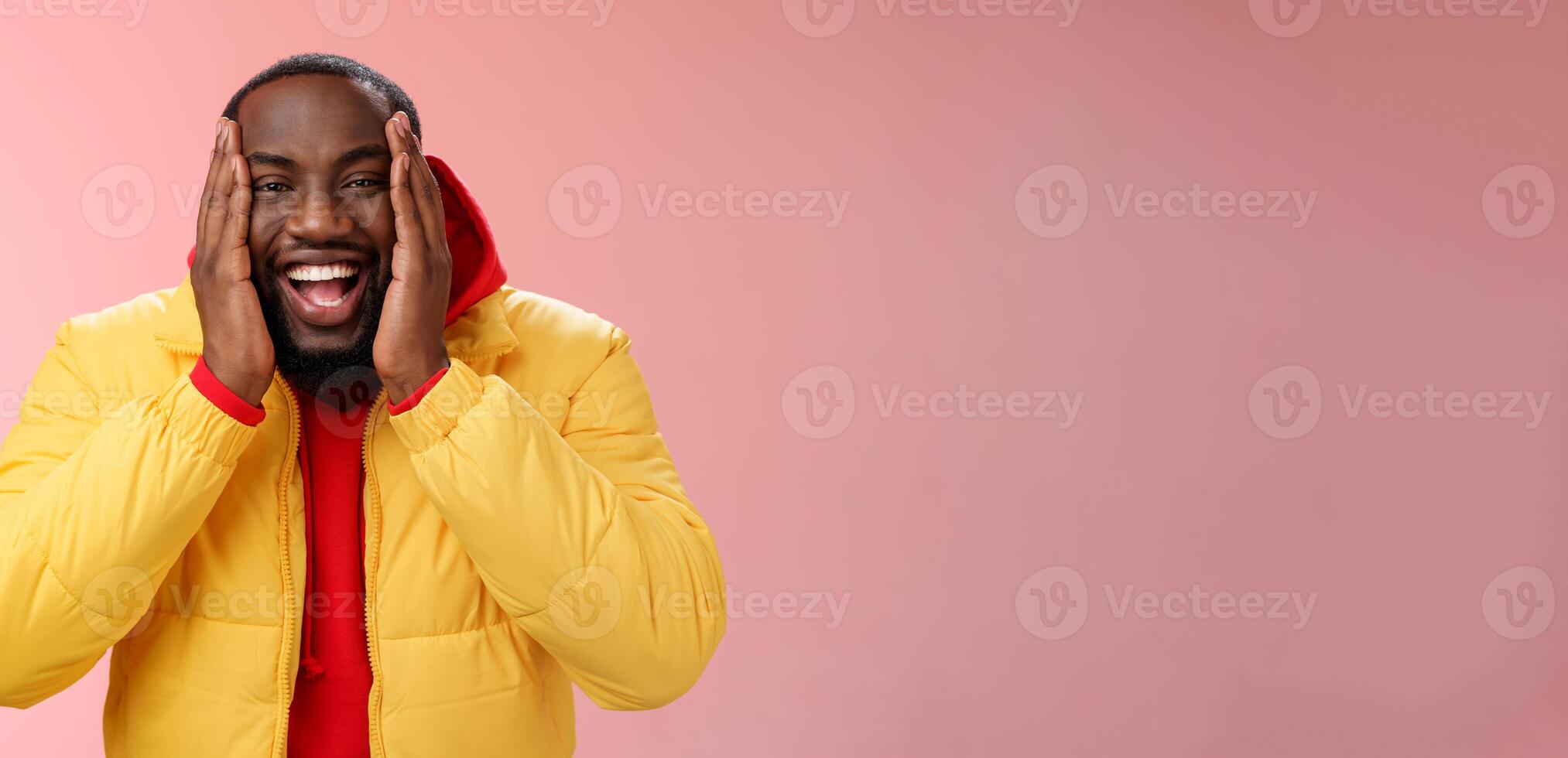 Happy charismatic playful african american bearded guy in yellow jacket red hoodie press palms cheeks smiling friendly laughing happily joking cannot believe receive awesome surprise, winning lottery photo