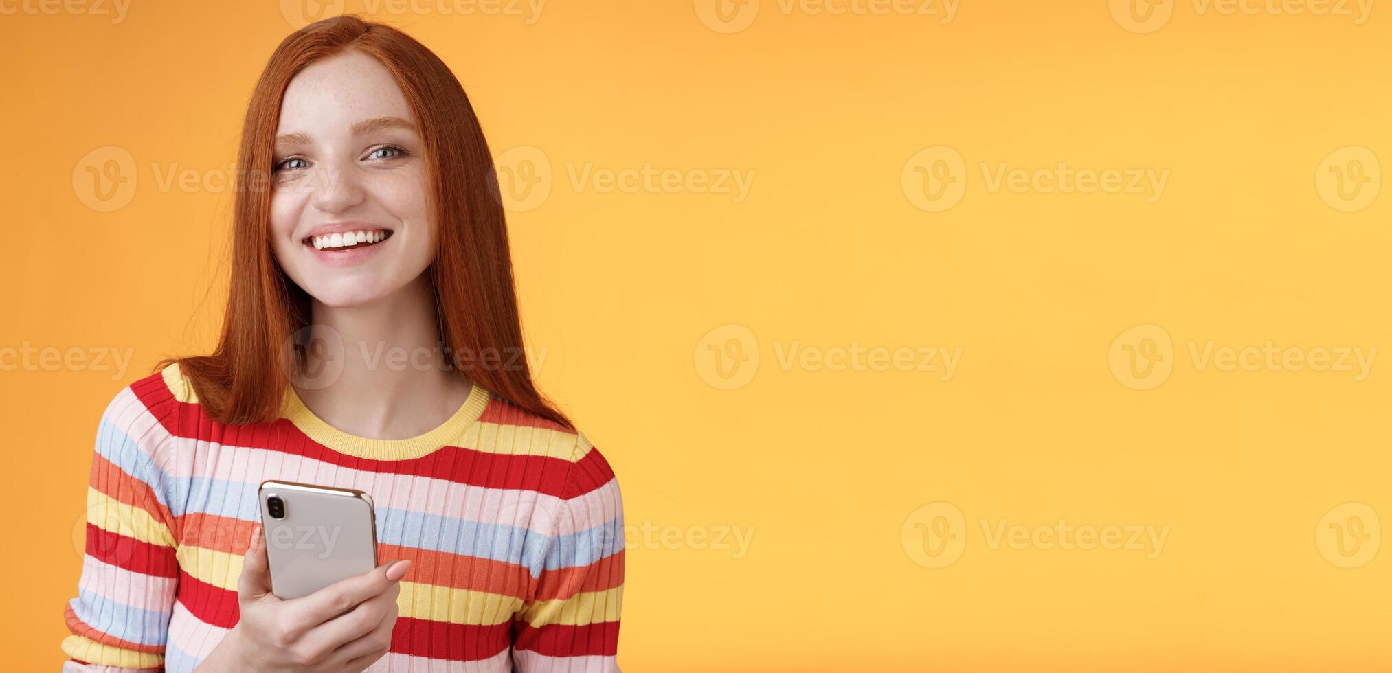 Friendly enthusiastic young redhead girl blue eyes using smartphone turn camera answer smiling broadly telling who sent message standing delighted orange background messaging, texting boyfriend photo