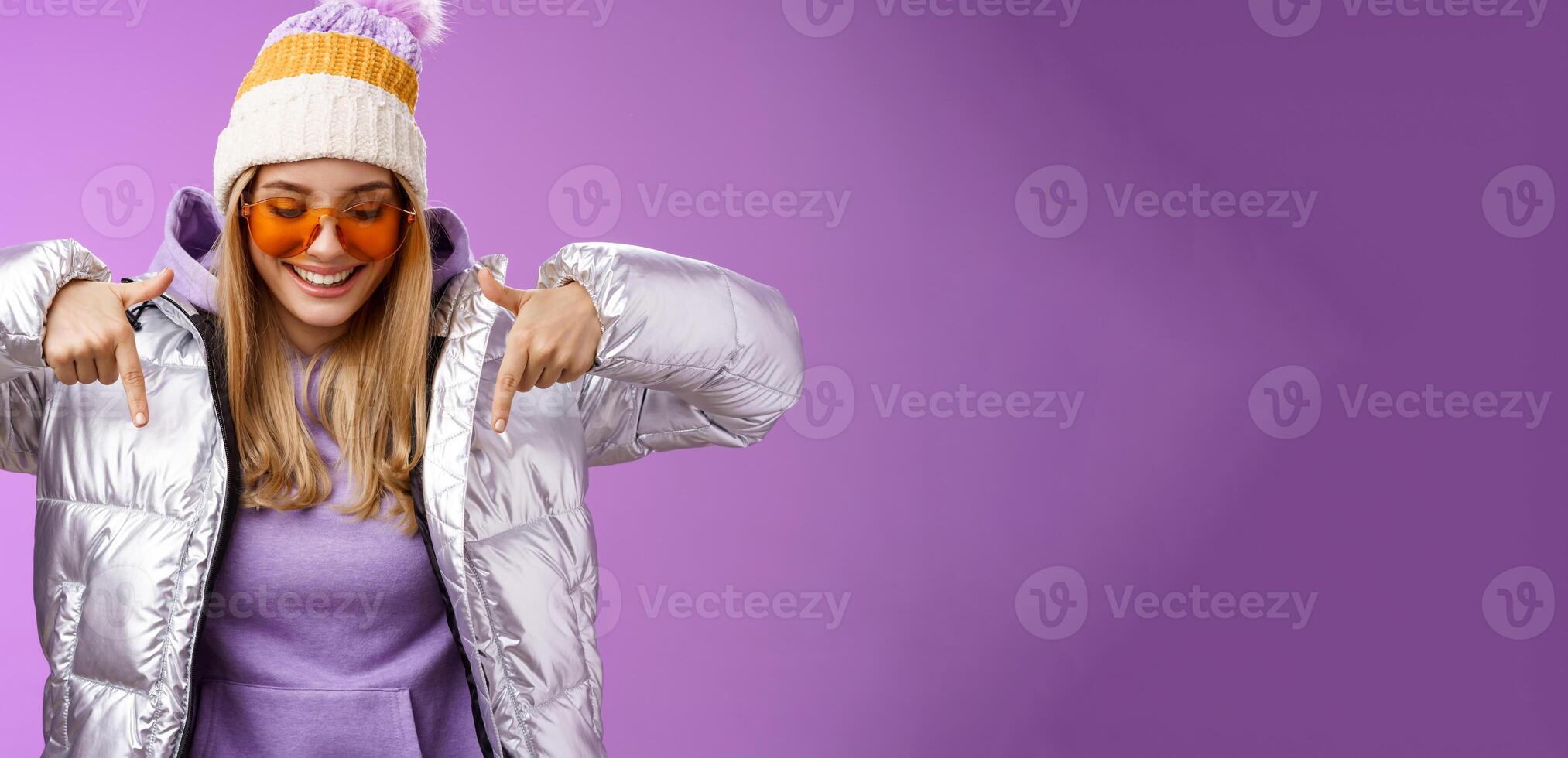 Joyful stylish blond woman in silver jacket stylish sunglasses jat enjoying perfect mountains view snowy vacation resort look pointing down smiling amused having fun feel happy, purple background photo