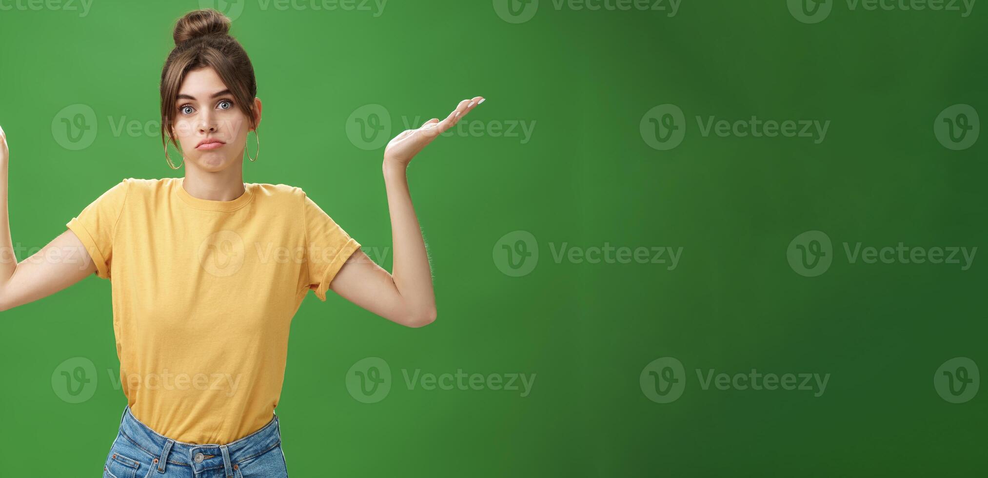 Girl do not know anything. Portrait of confused unsure cute woman with combed hair shrugging with raised hands pouting looking clueless at camera standing questioned against green background photo